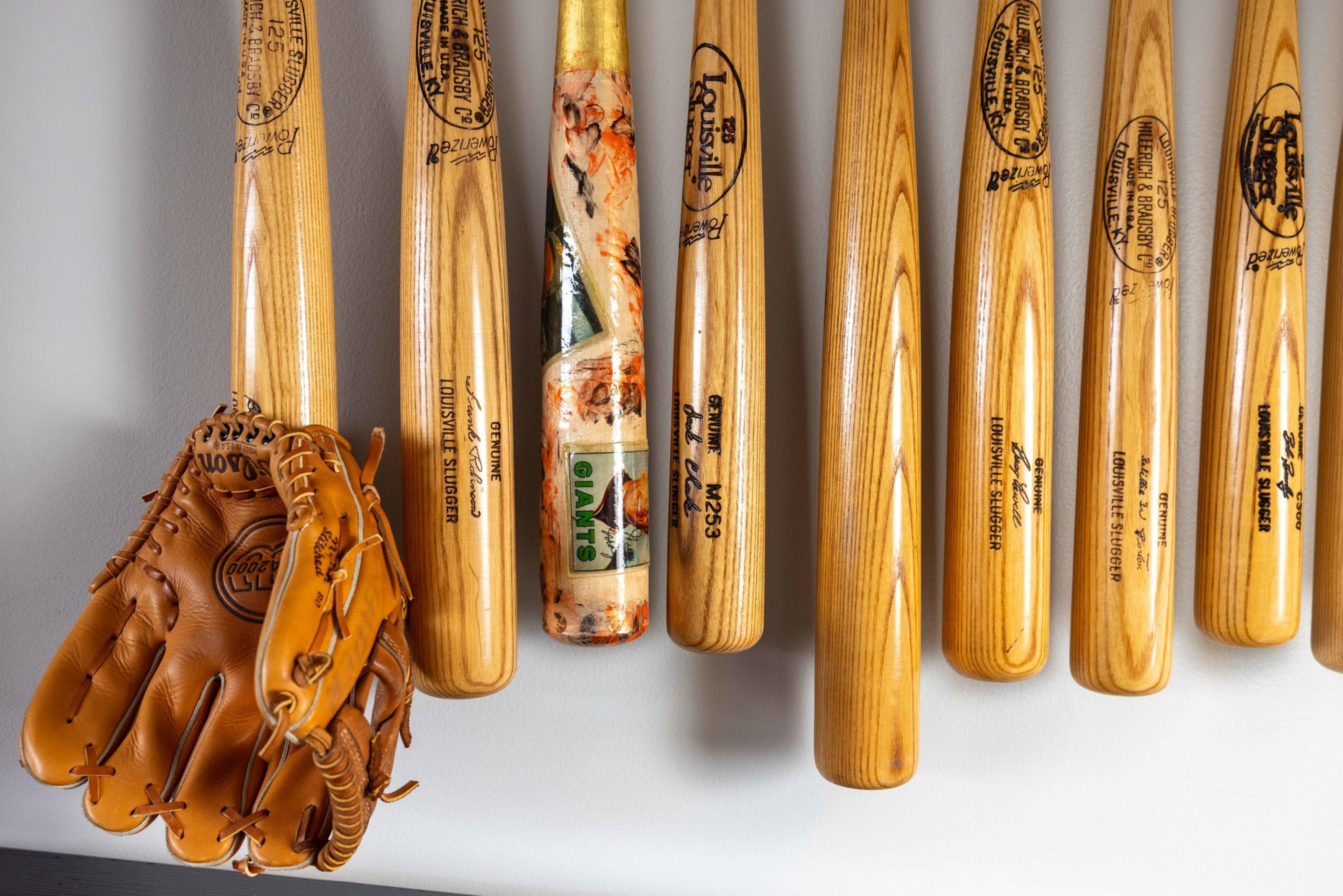 Several wooden baseball bats are hanging vertically on a wall, with a pair of brown leather baseball gloves resting on one of the bats.