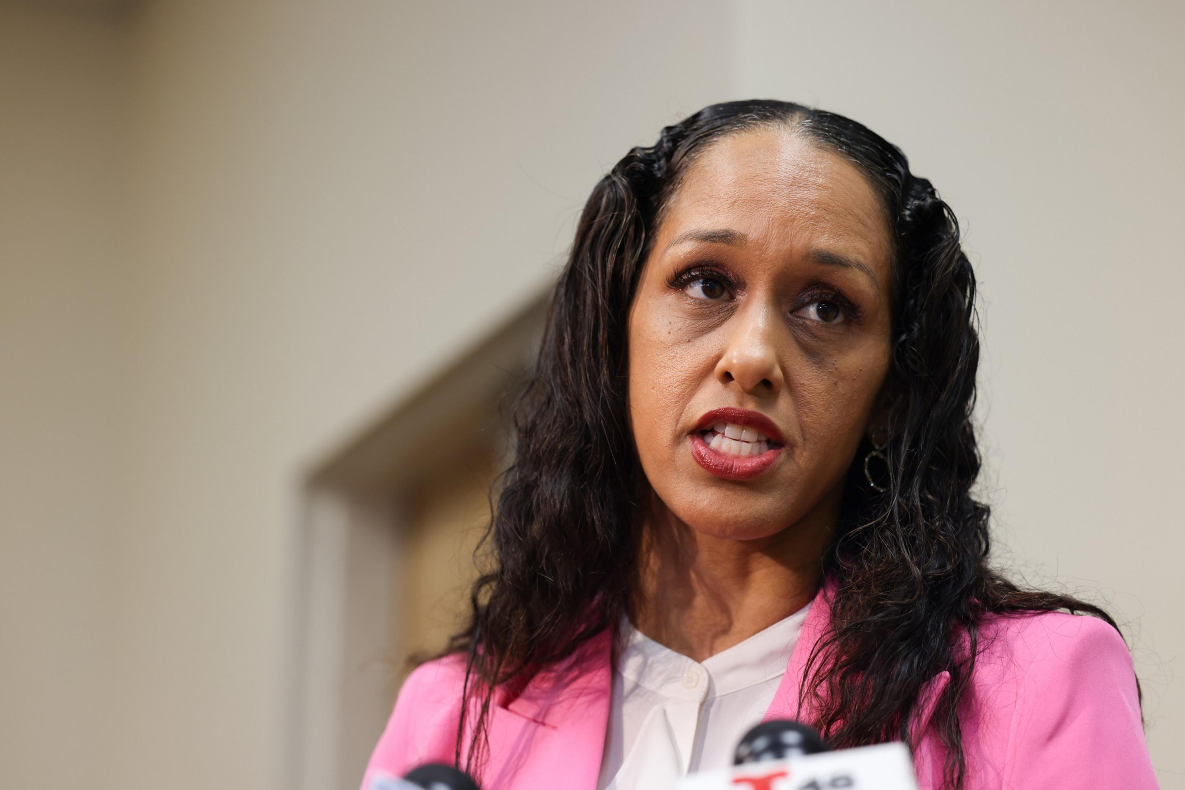 A woman with long, curly hair, wearing a pink blazer and white blouse, speaks in front of multiple microphones. The background is plain, with a blurred doorway.