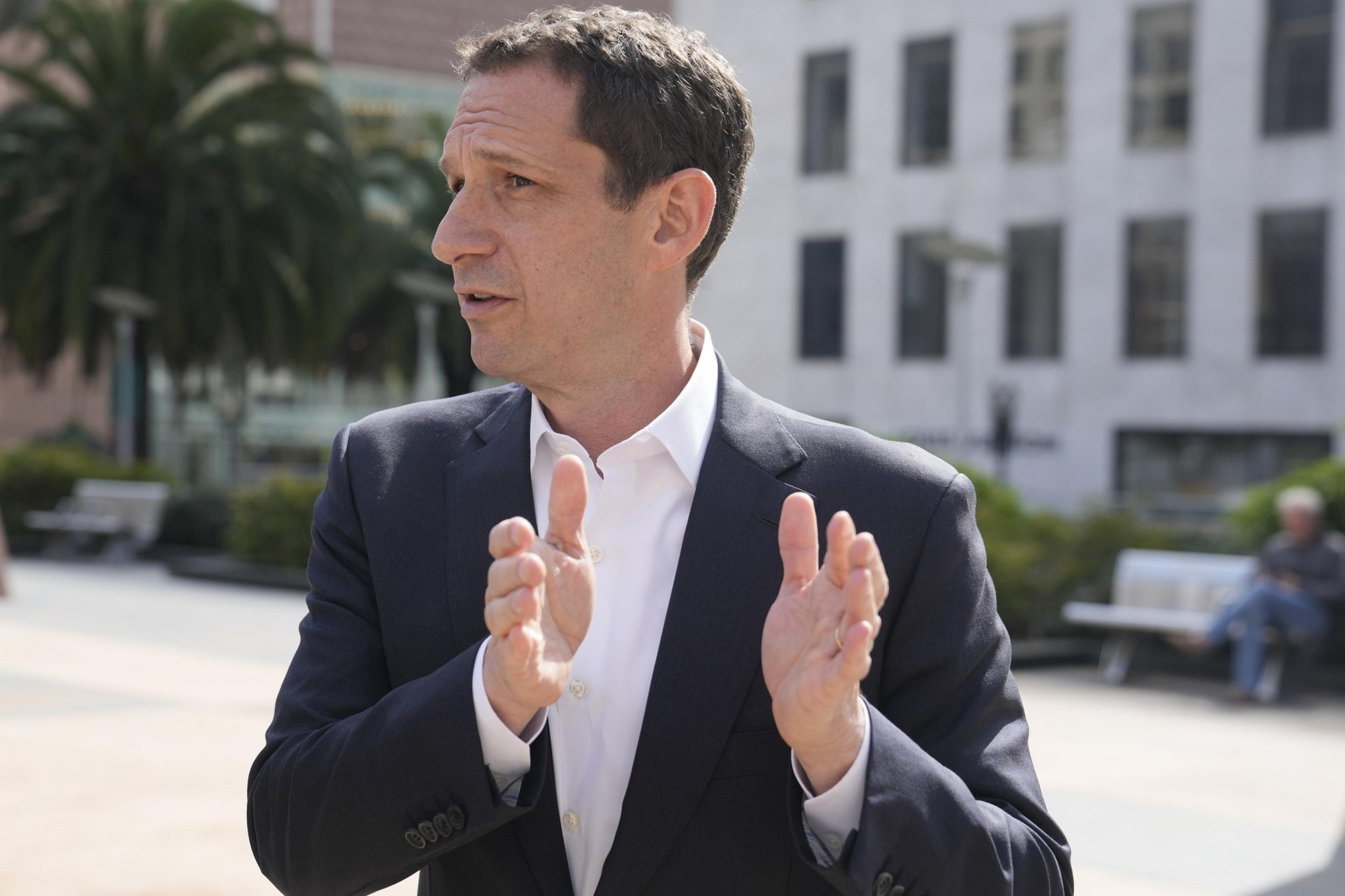 A person in a suit gestures outdoors in a city setting with palm trees and buildings in the background, conveying a sense of discussion or explanation.