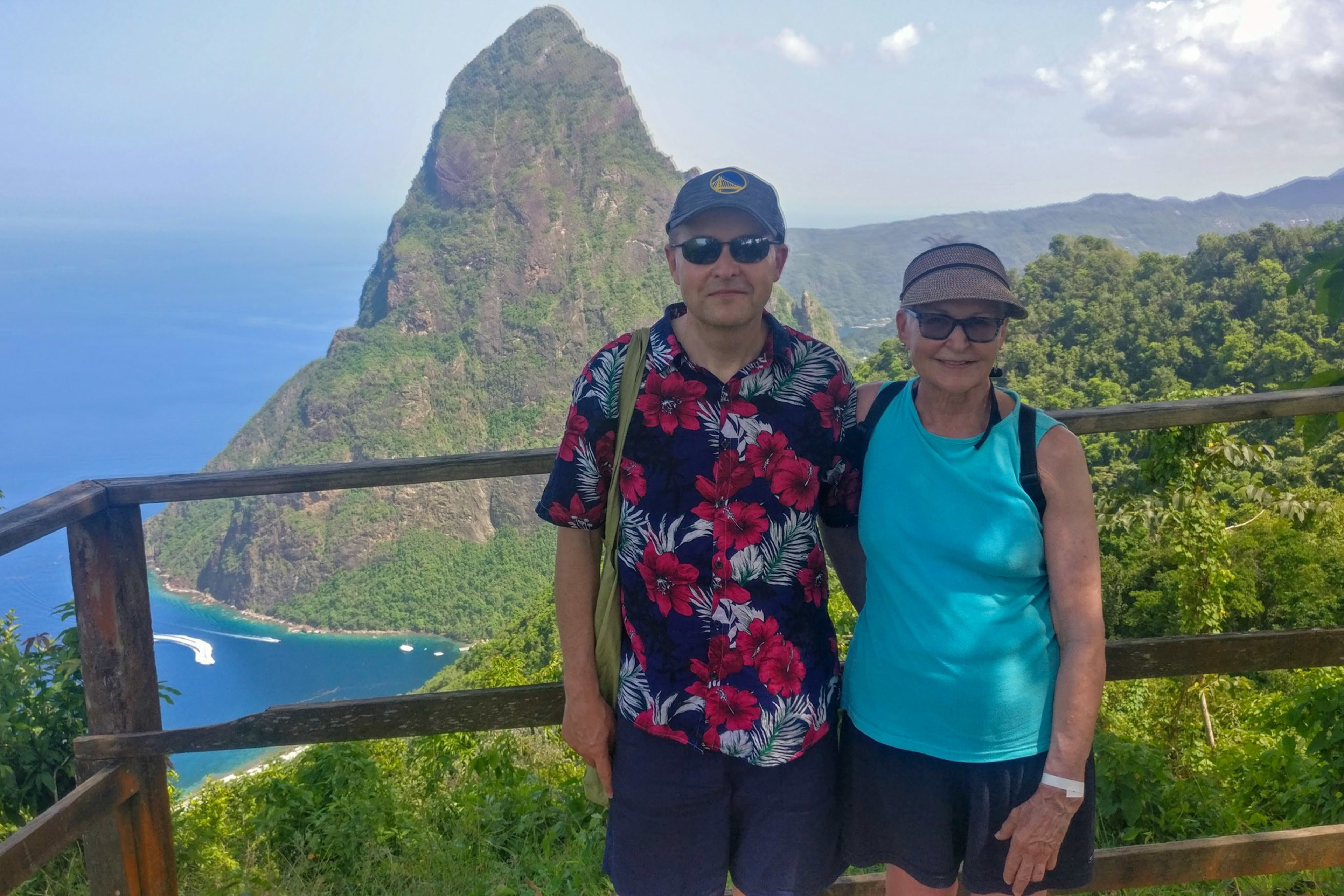 A man and woman wearing sunglasses stand together on a wooden balcony with a tall, pointed mountain and blue ocean in the background.