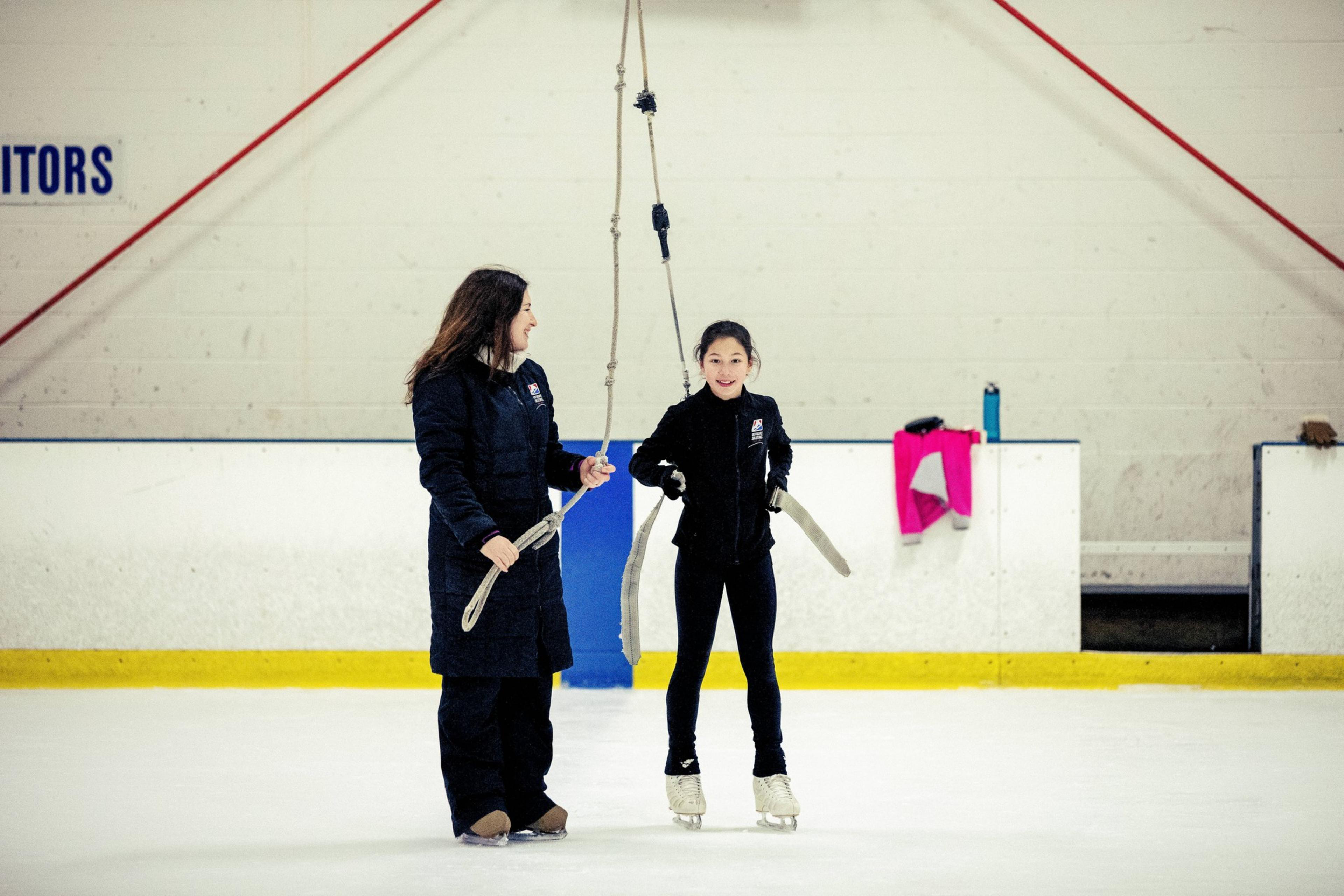 A skating instructor helps a young girl on ice skates hold a support strap at an indoor ice rink.
