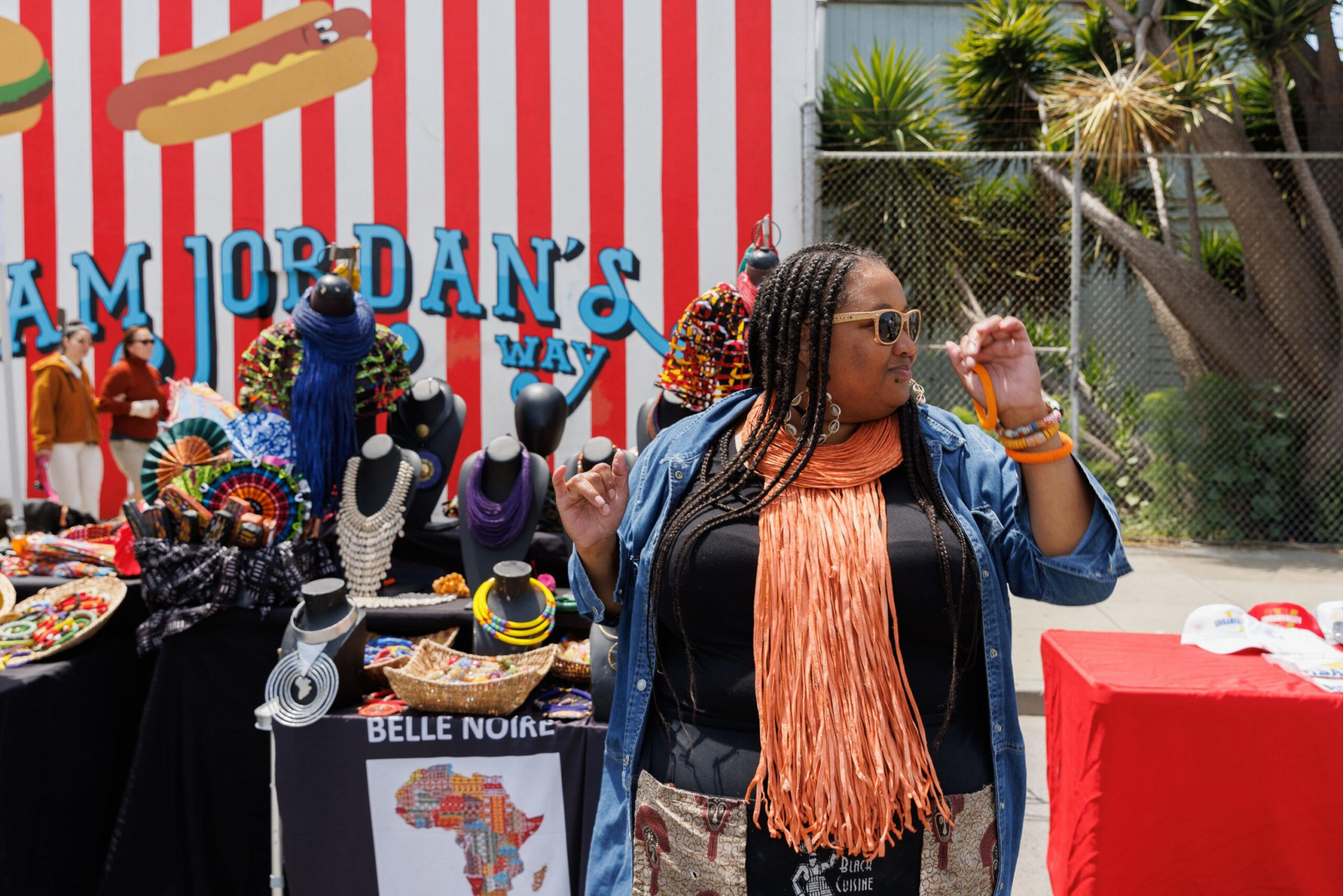 A woman in sunglasses stands near a table displaying colorful crafts, with a red-striped background behind her.