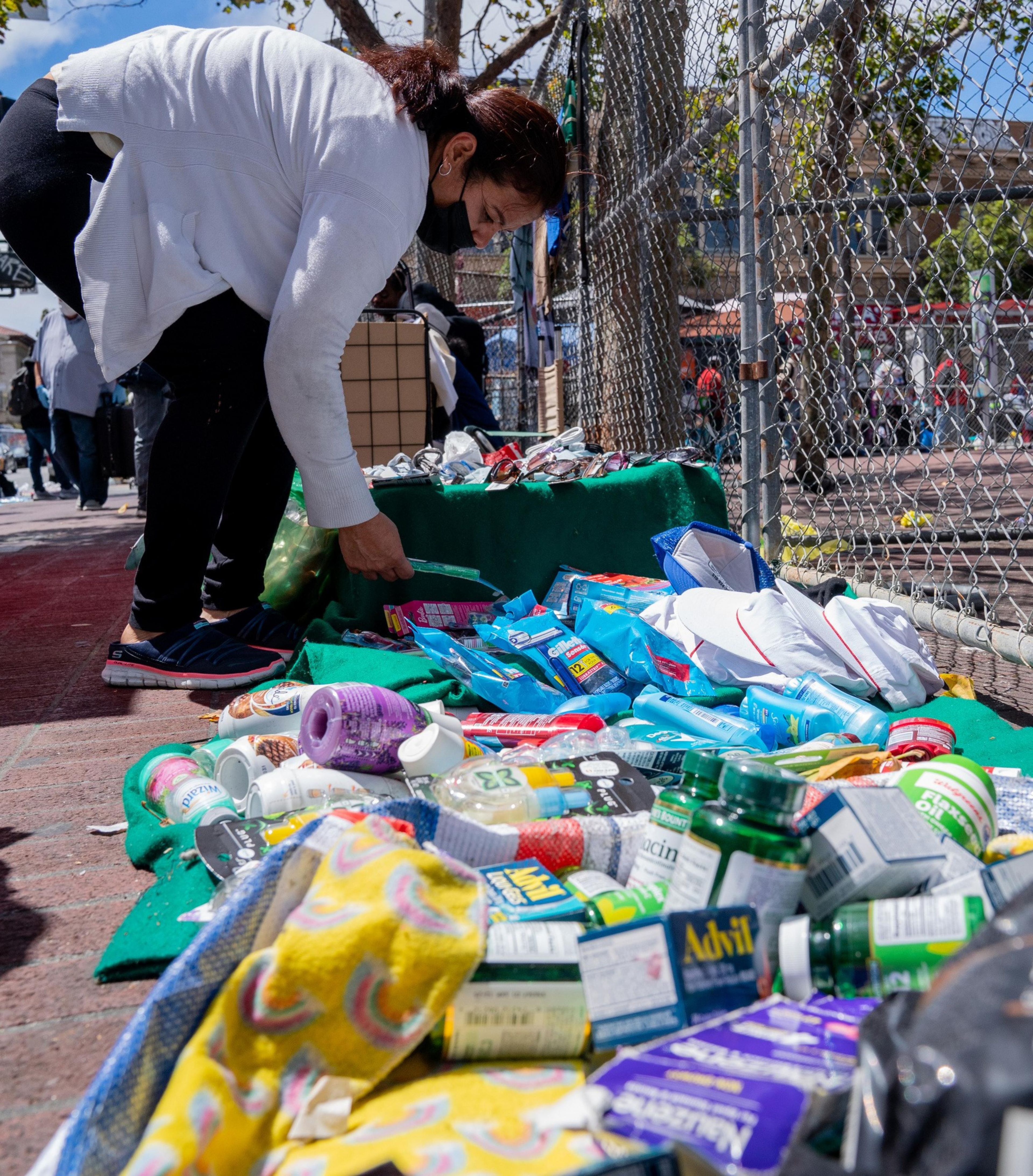 A person in a white jacket and black mask browses a sidewalk display of various toiletry items and medicine packages, spread on a green cloth near a chain-link fence.