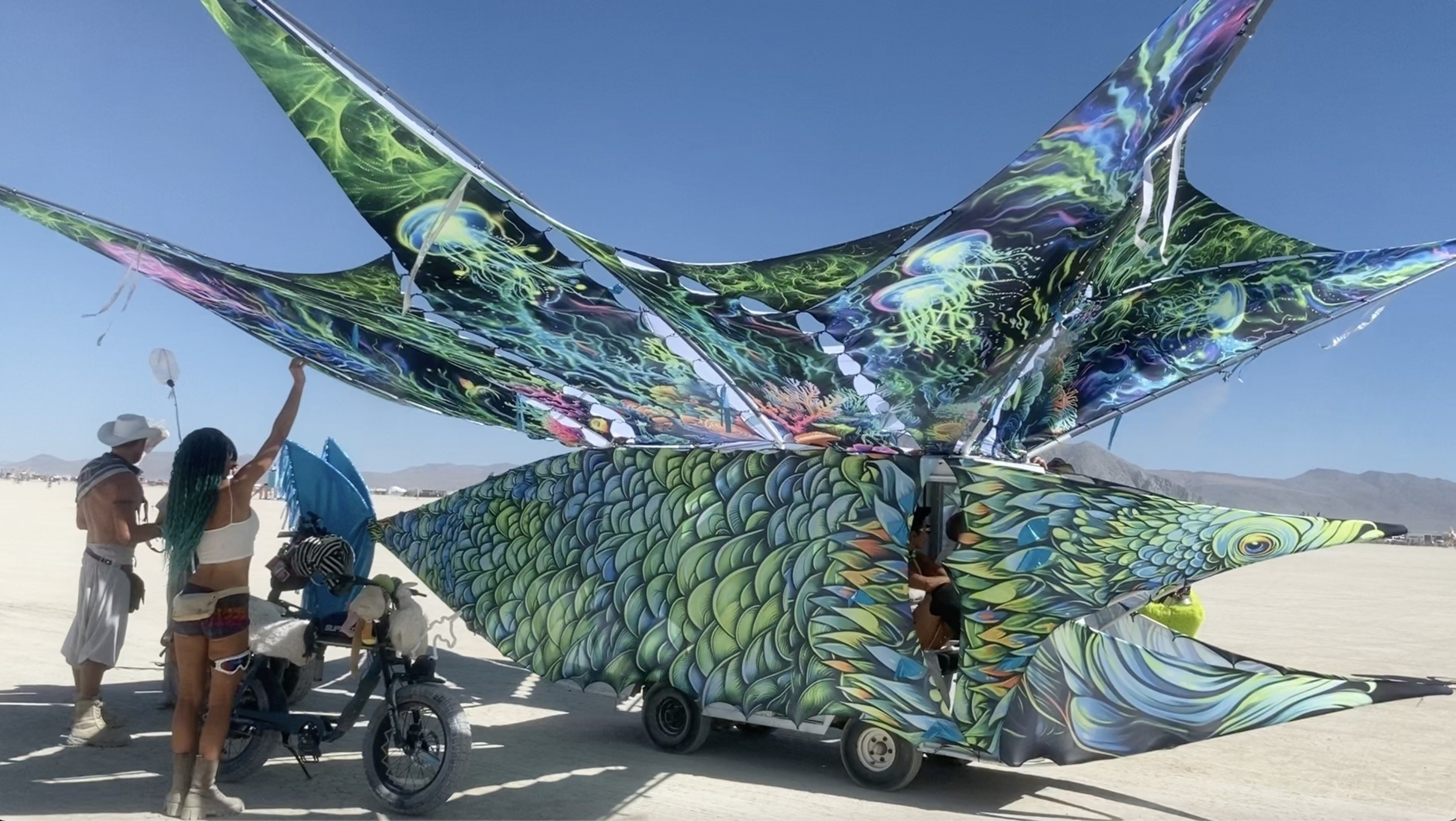 A colorful fish-shaped vehicle with large, vibrant fins and scales is parked on a desert plain with three people nearby under a clear blue sky.