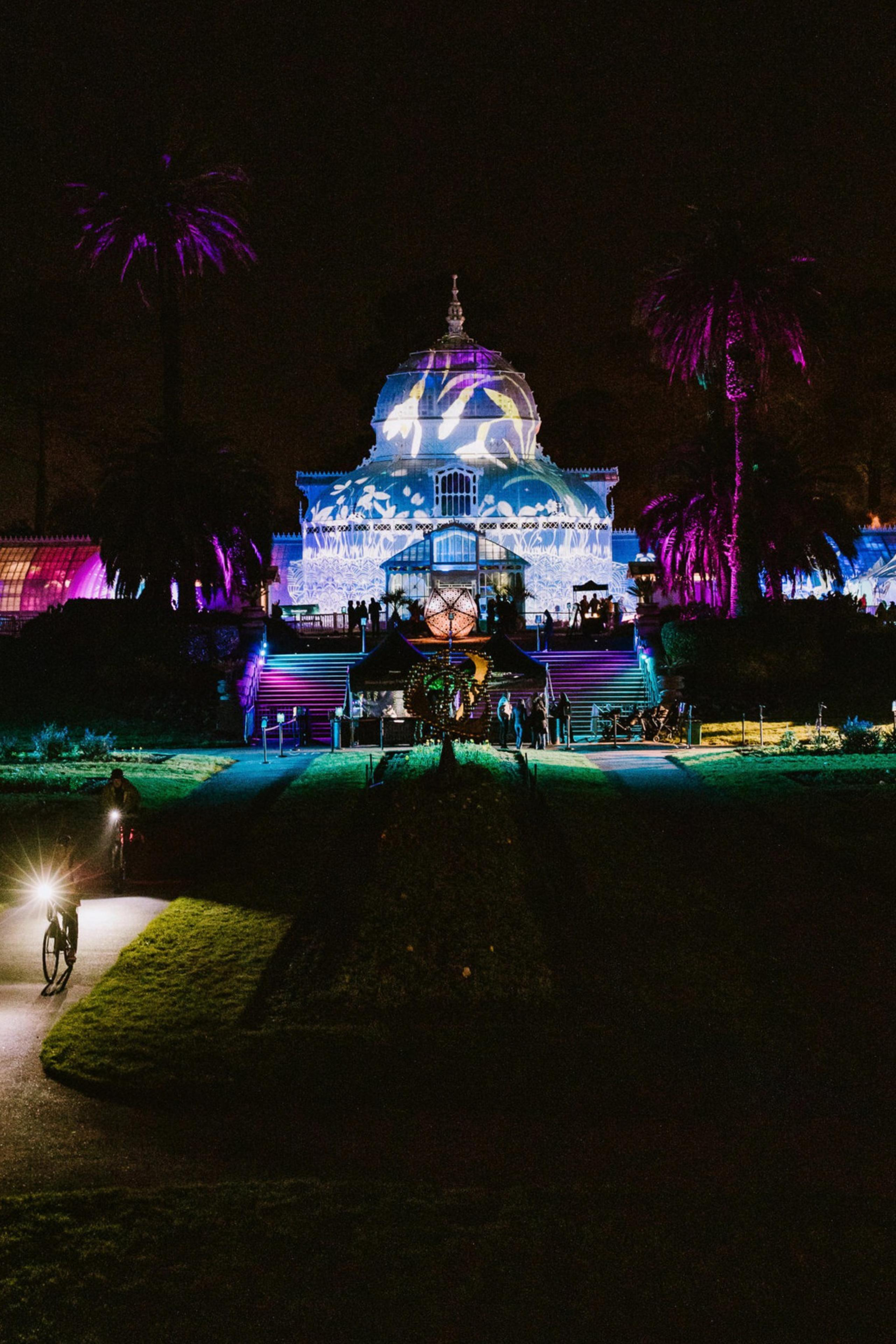 A grand, white, dome-shaped building is illuminated with colorful lights and patterns, set against a backdrop of palm trees, while people and bikers pass by at night.