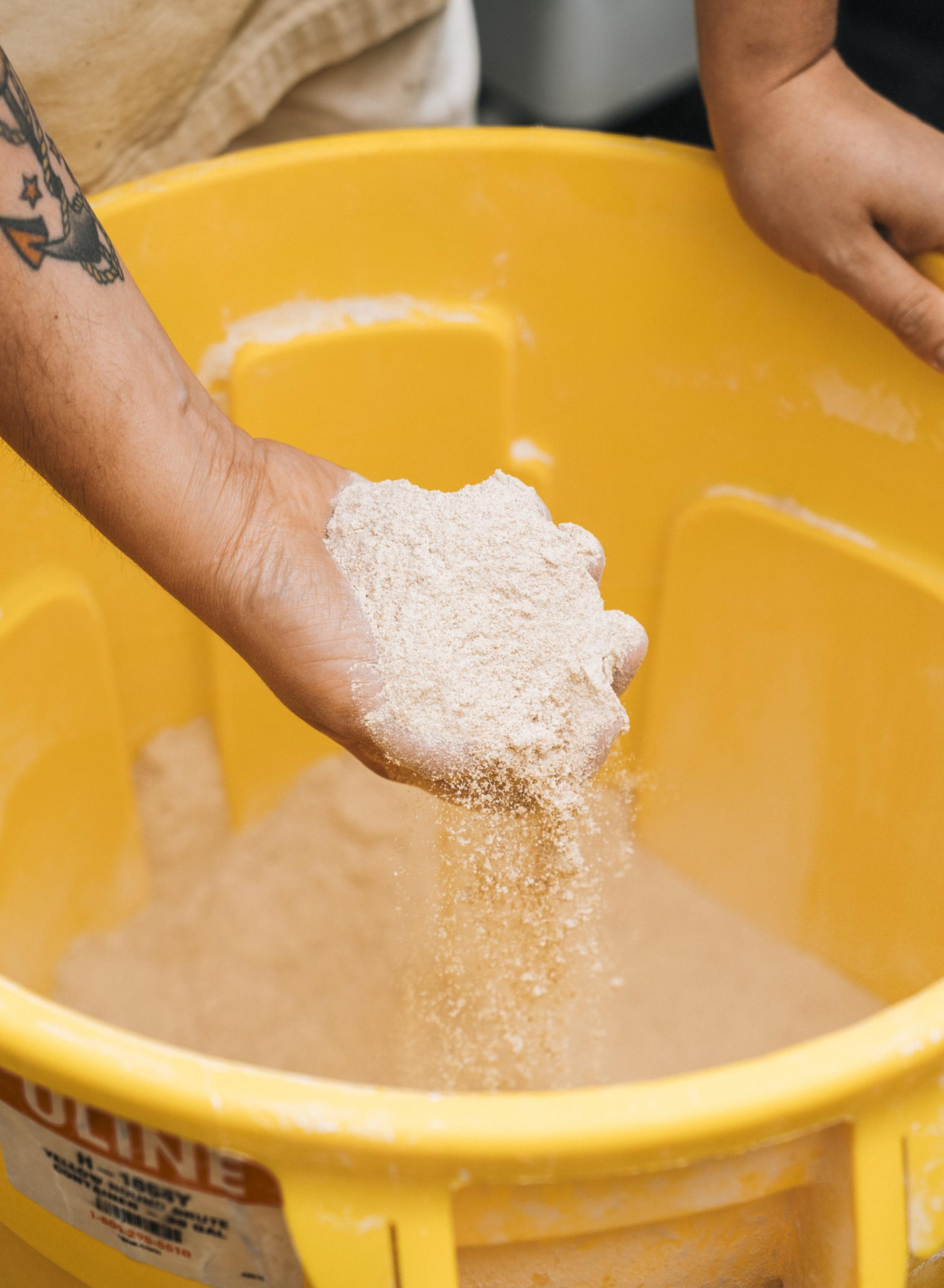 A hand holds a handful of flour, allowing it to fall into a large yellow container. The arm has a tattoo and another hand rests on the container's edge.