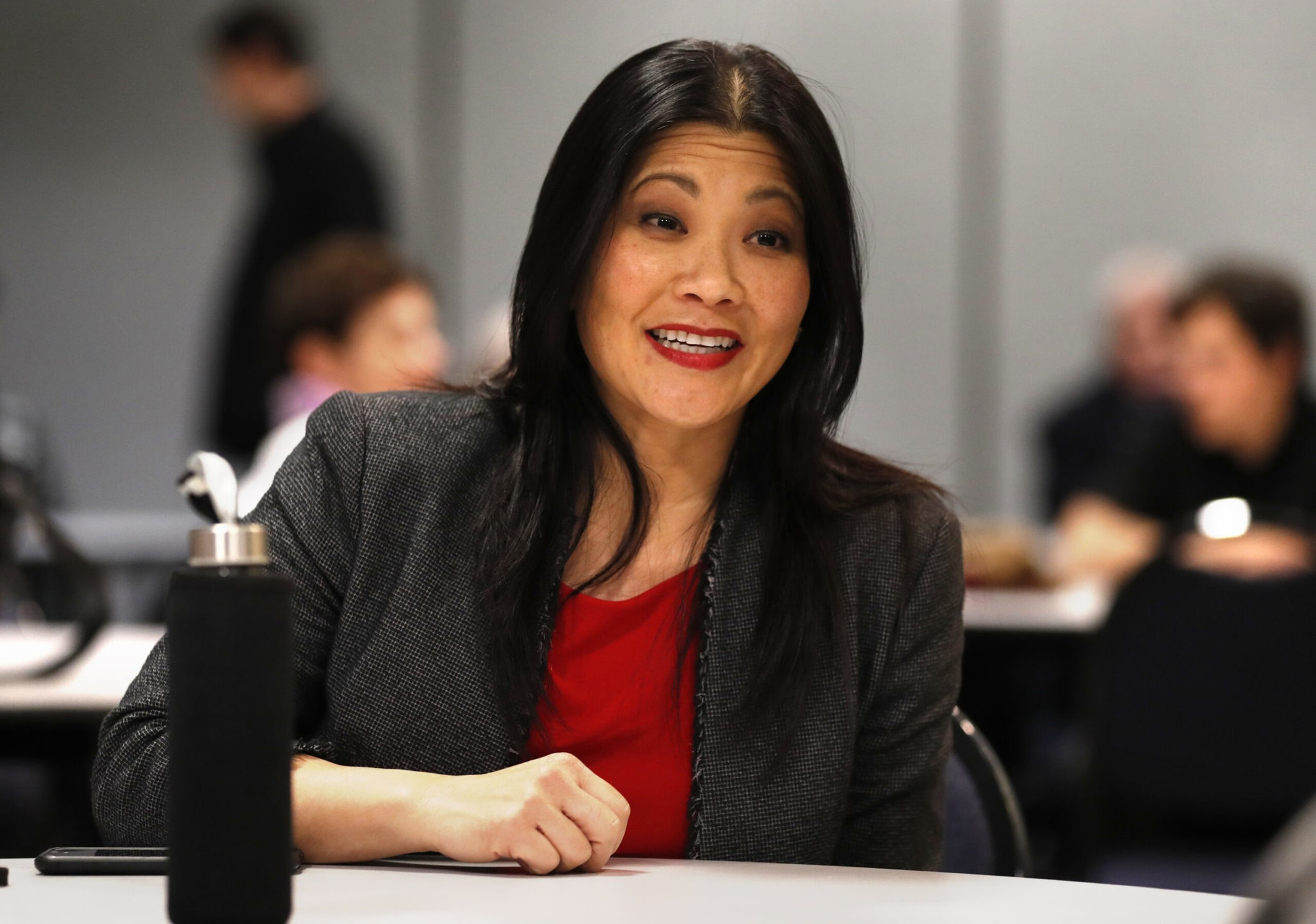 A woman with long black hair, wearing a gray blazer and red top, sits at a table smiling, with a black water bottle in front of her.