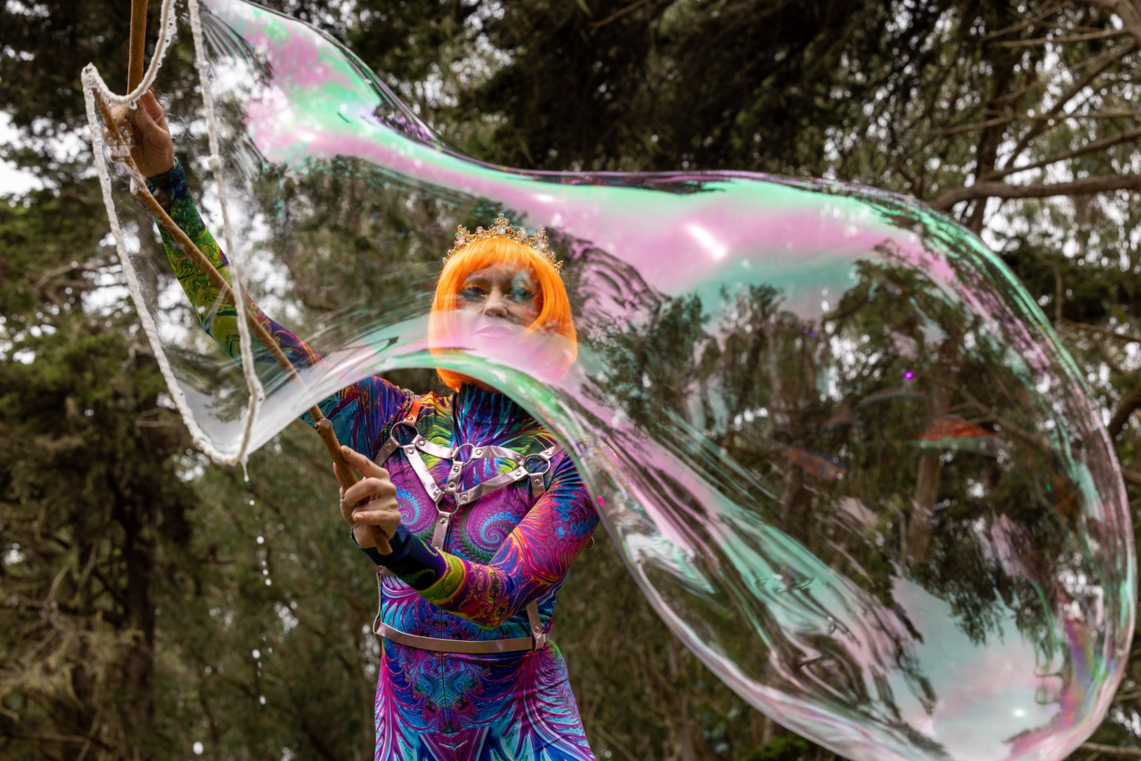 A person with bright orange hair and colorful, patterned clothes is creating a giant iridescent soap bubble outdoors among trees.