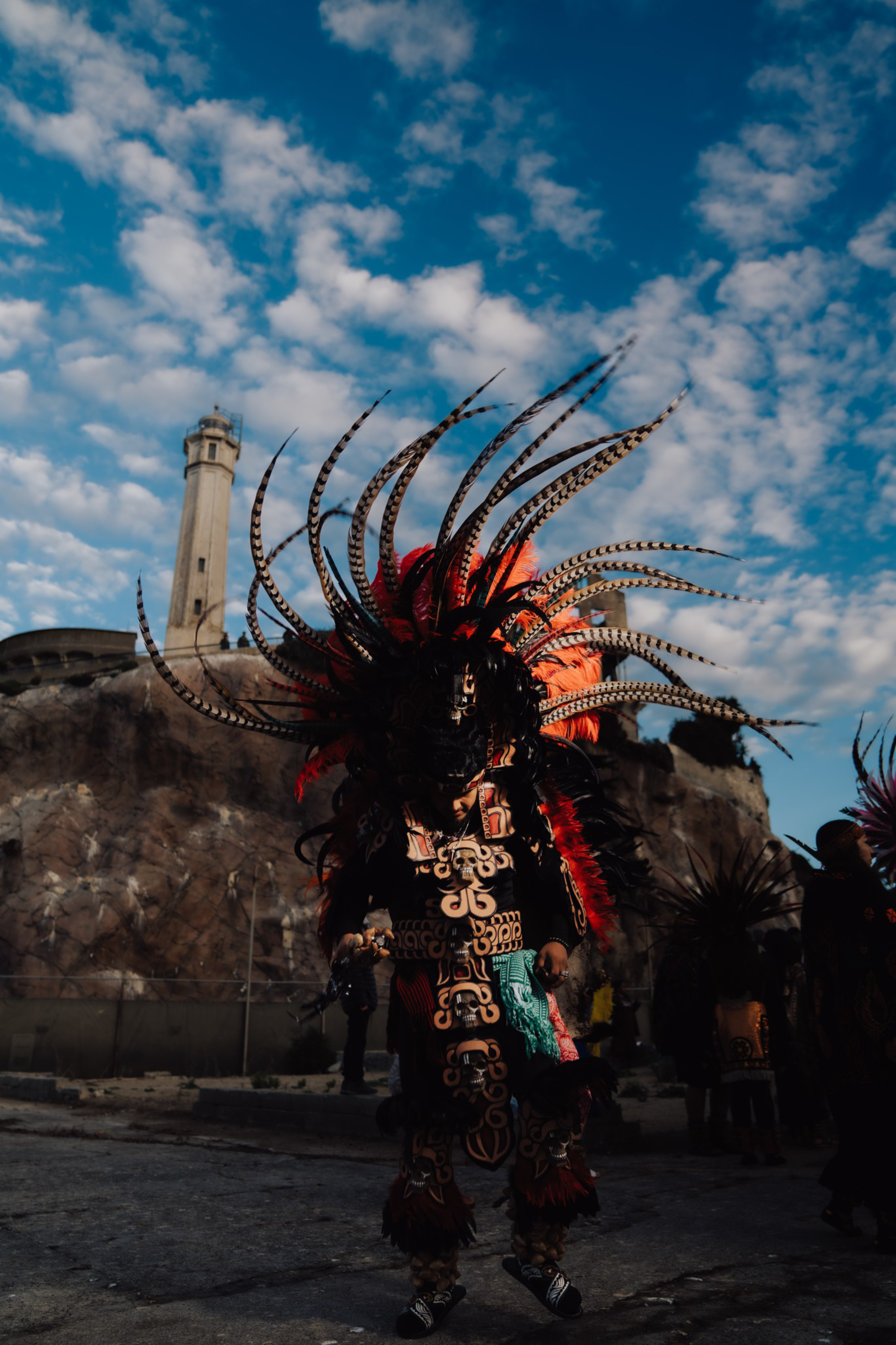 A person wears an elaborate Aztec warrior costume with tall feathered headdress and ornate patterns, standing in front of a lighthouse against a partly cloudy sky.