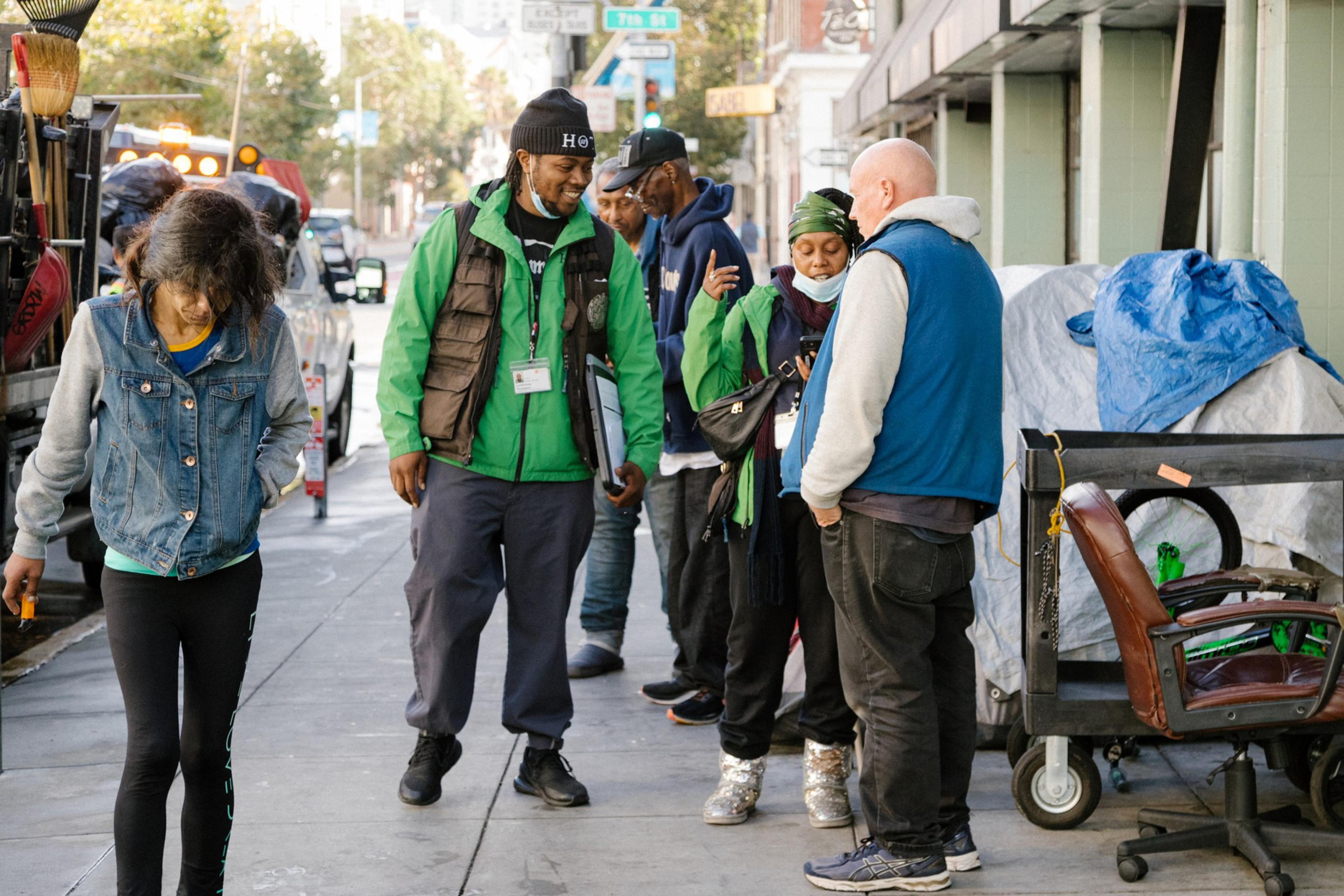 A group of people stands on a city sidewalk, some in conversation. One person walks while others are near a cart with a chair and covered items.