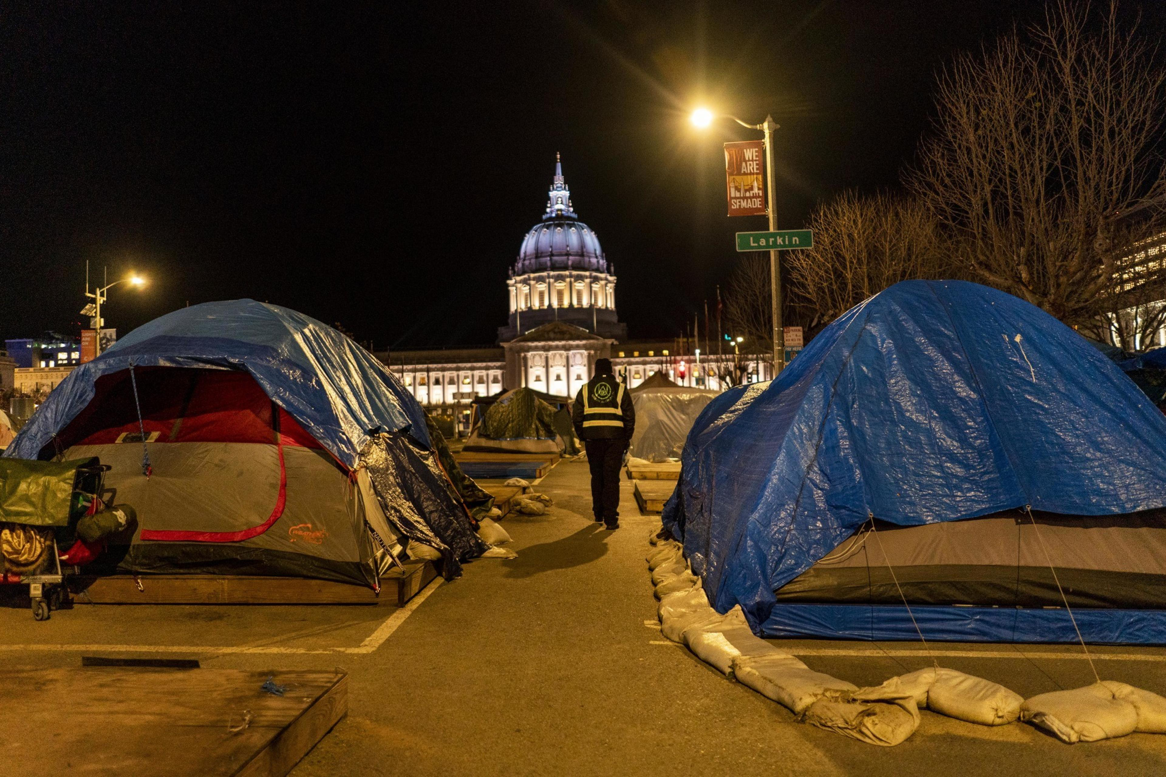 A row of tents covered with blue tarps is set up on a street at night. A person in a vest walks by, with a domed, illuminated building in the background.
