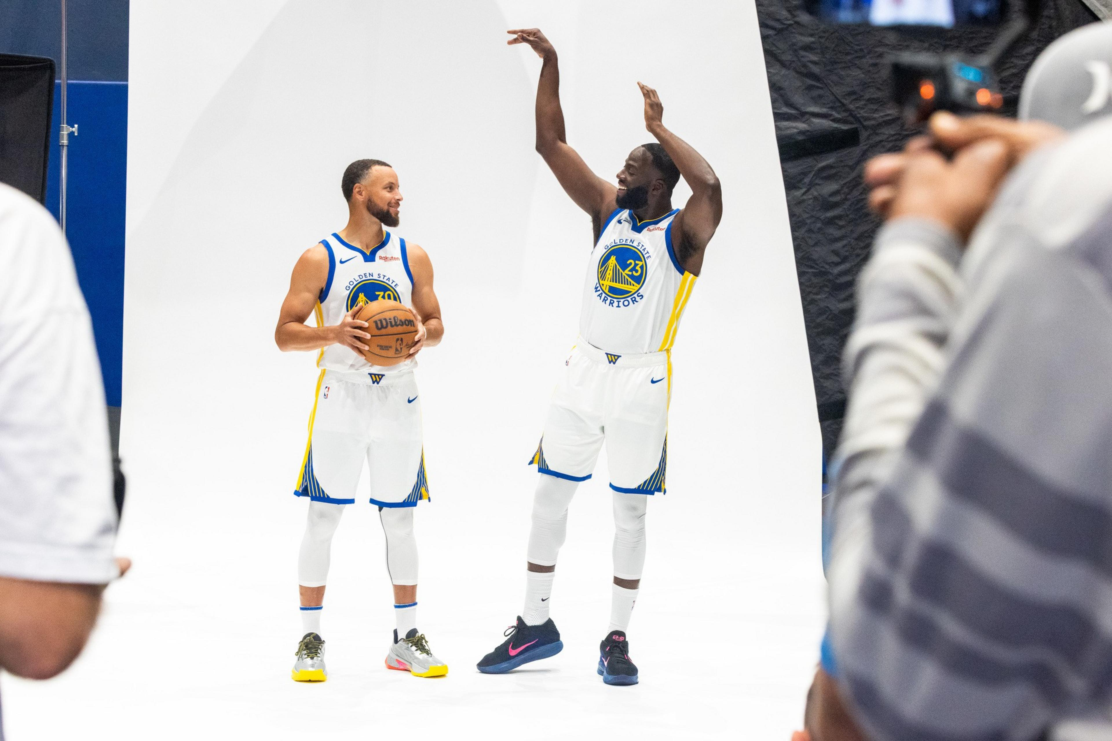 Two Golden State Warriors players pose for photos on a white backdrop, one holding a basketball while the other raises his arms as if shooting.