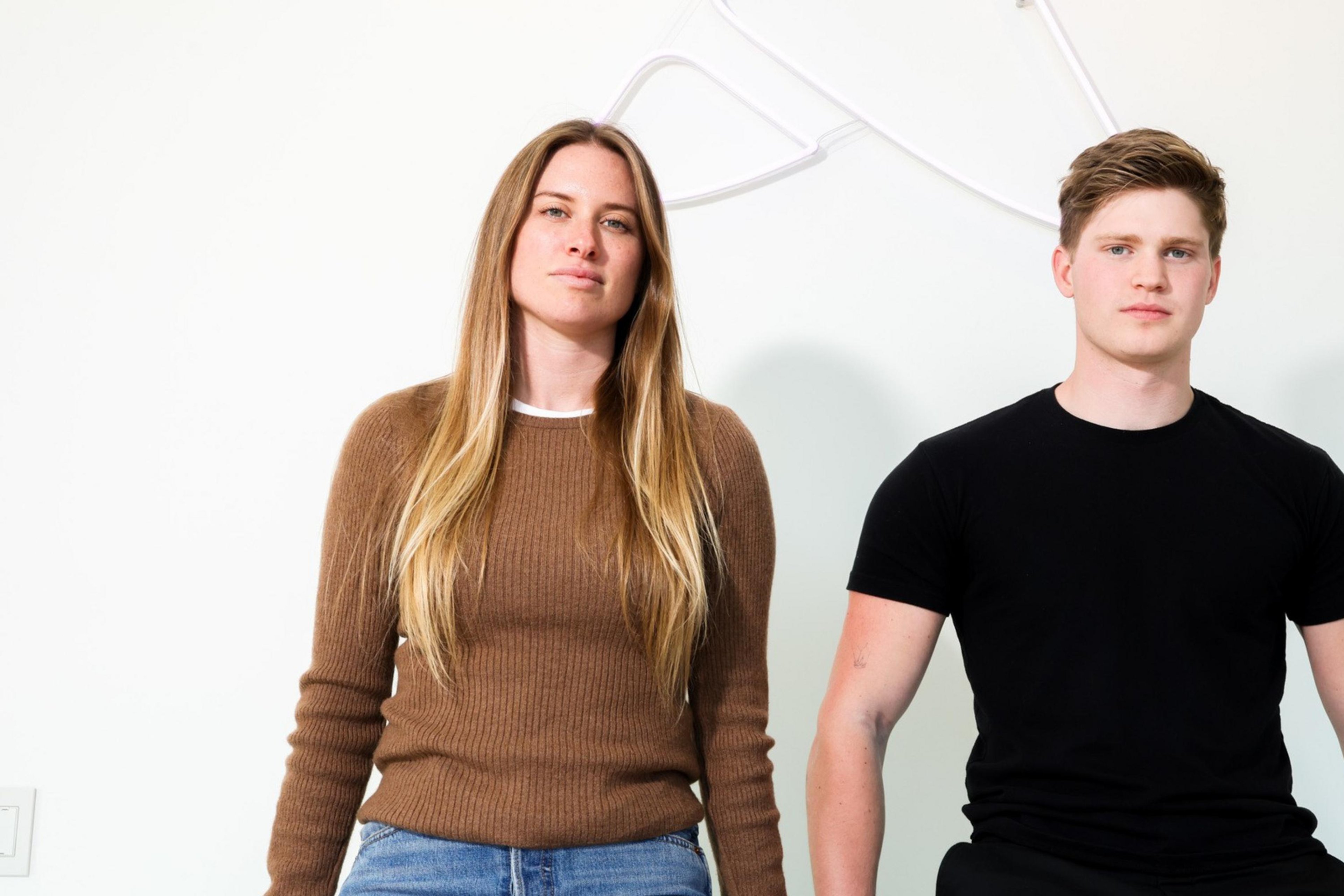 A woman in a brown sweater and jeans and a man in a black t-shirt and pants sit casually on a white surface against a plain background.