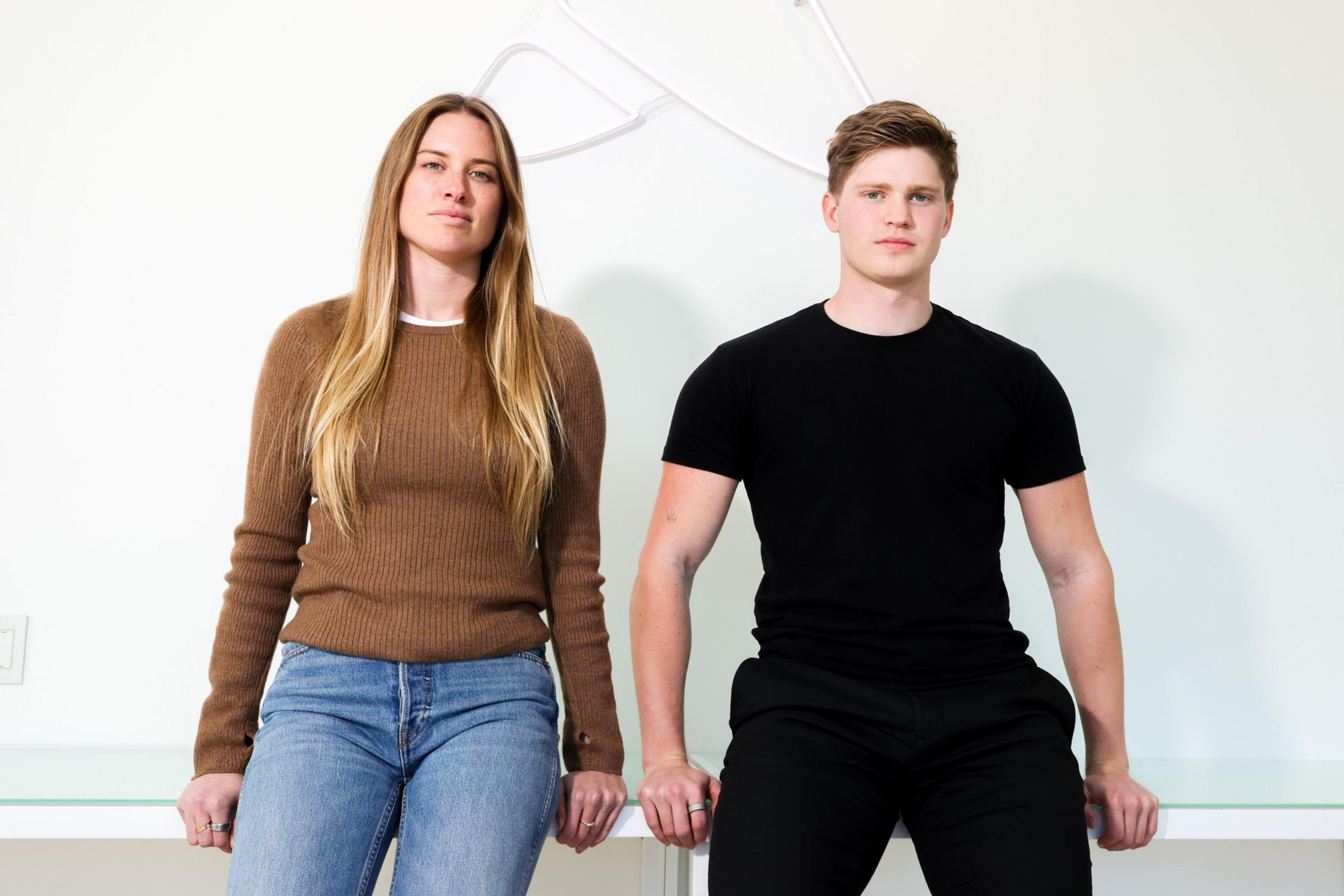 A woman in a brown sweater and jeans and a man in a black t-shirt and pants sit casually on a white surface against a plain background.