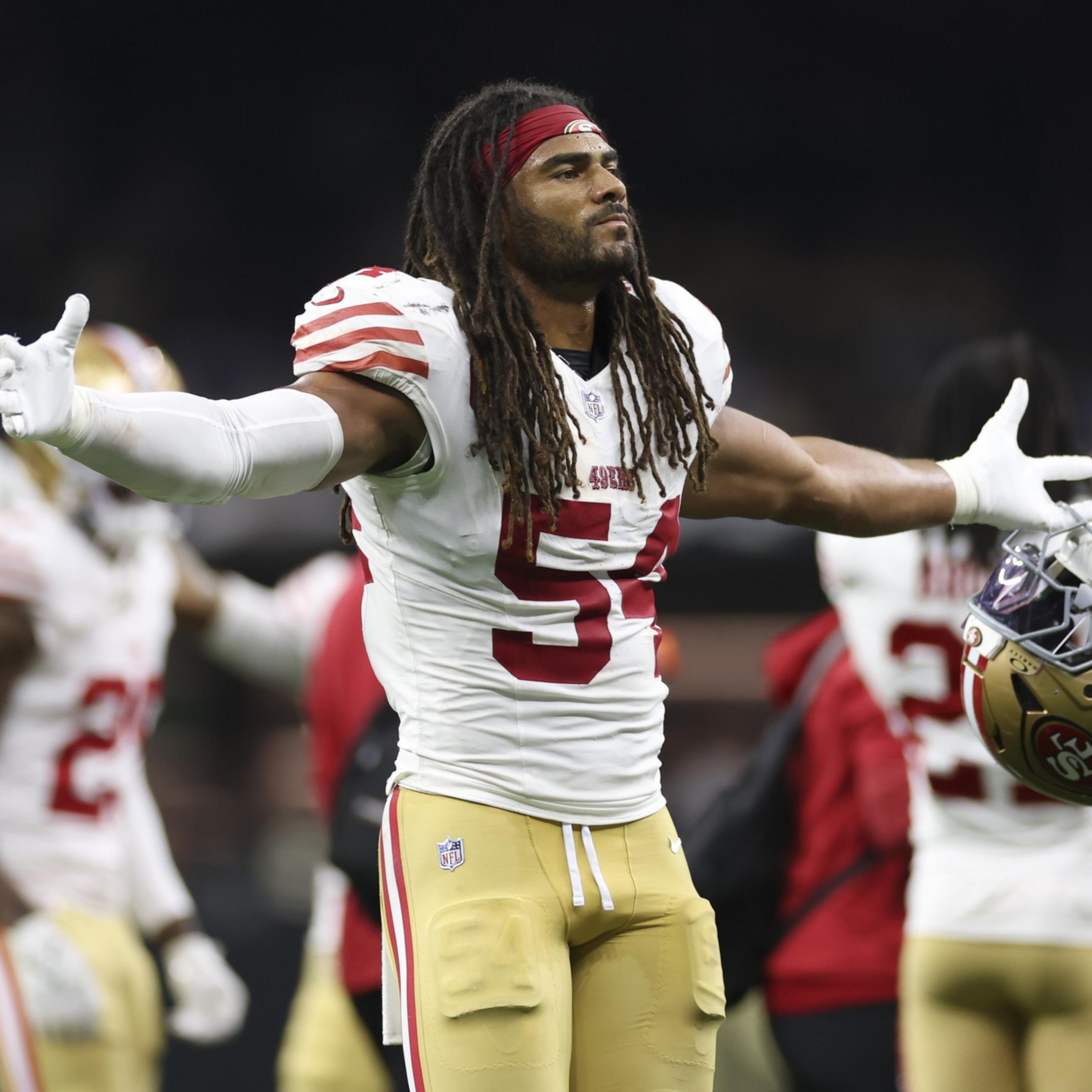 A football player with dreadlocks and a red headband, wearing a white and red jersey with number 54, holds his helmet in one hand and spreads his arms wide.