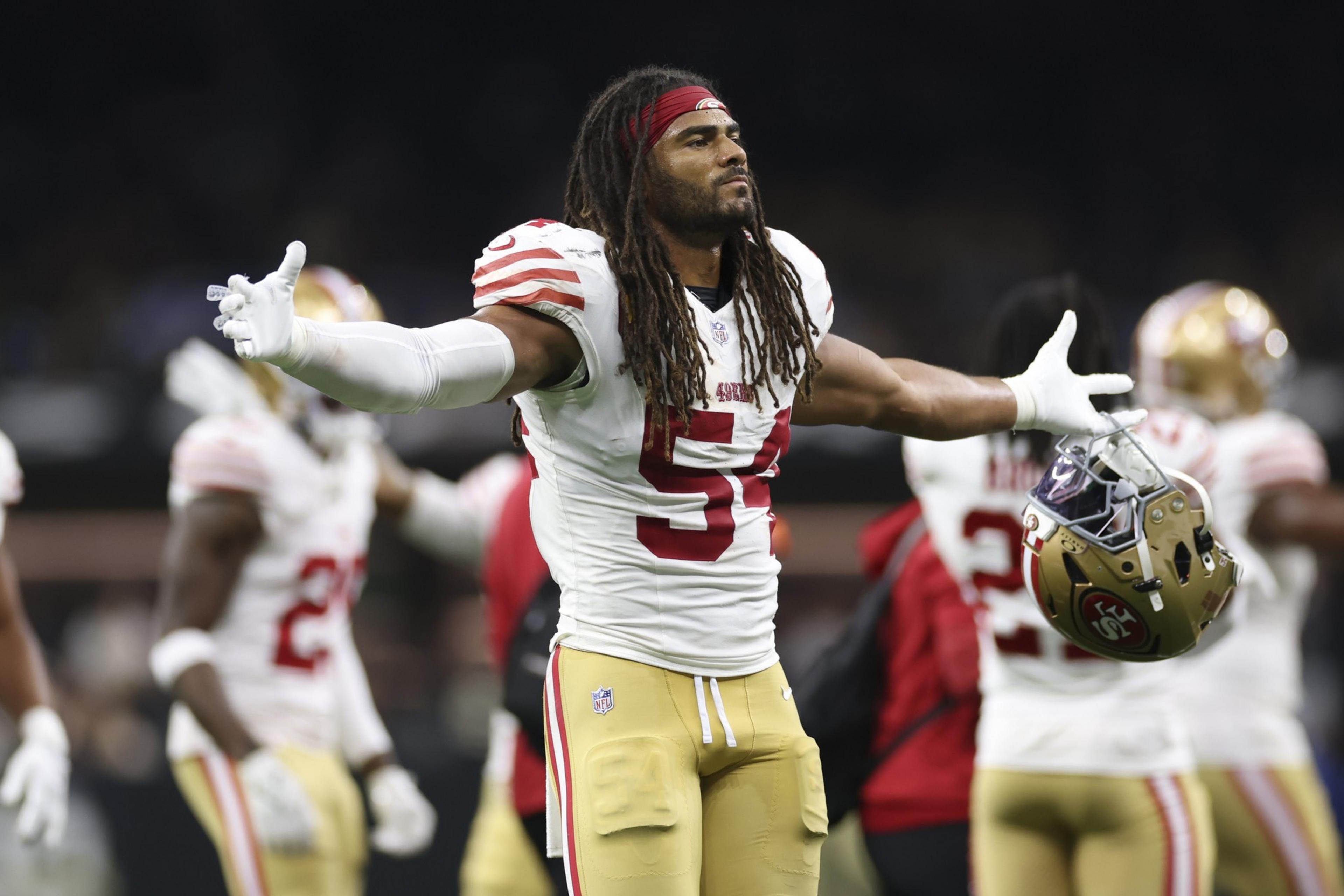 A football player with dreadlocks and a red headband, wearing a white and red jersey with number 54, holds his helmet in one hand and spreads his arms wide.