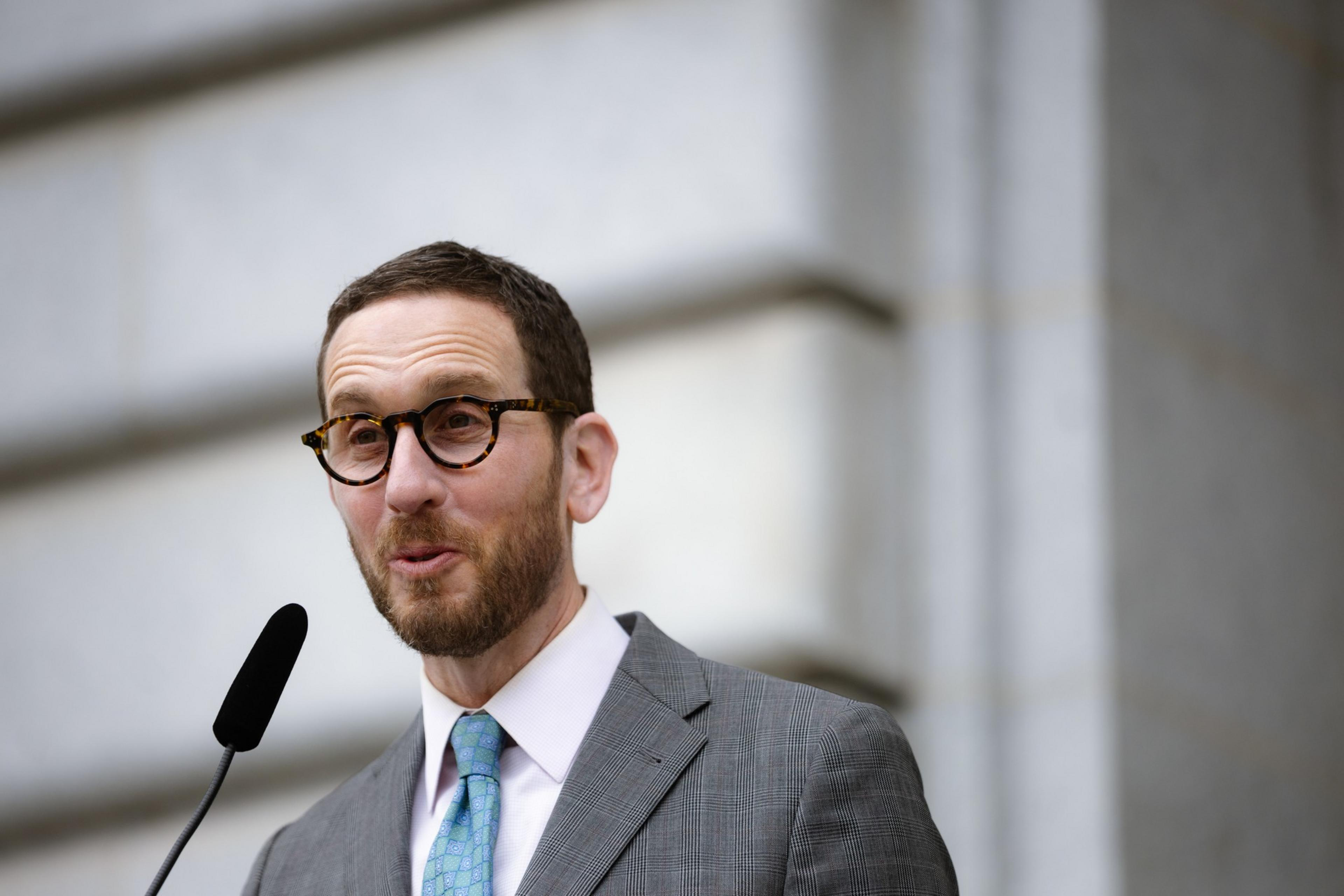 A man in a gray suit and blue tie speaks into a microphone. He has short hair, a beard, and is wearing round glasses. The background is a blurred stone wall.