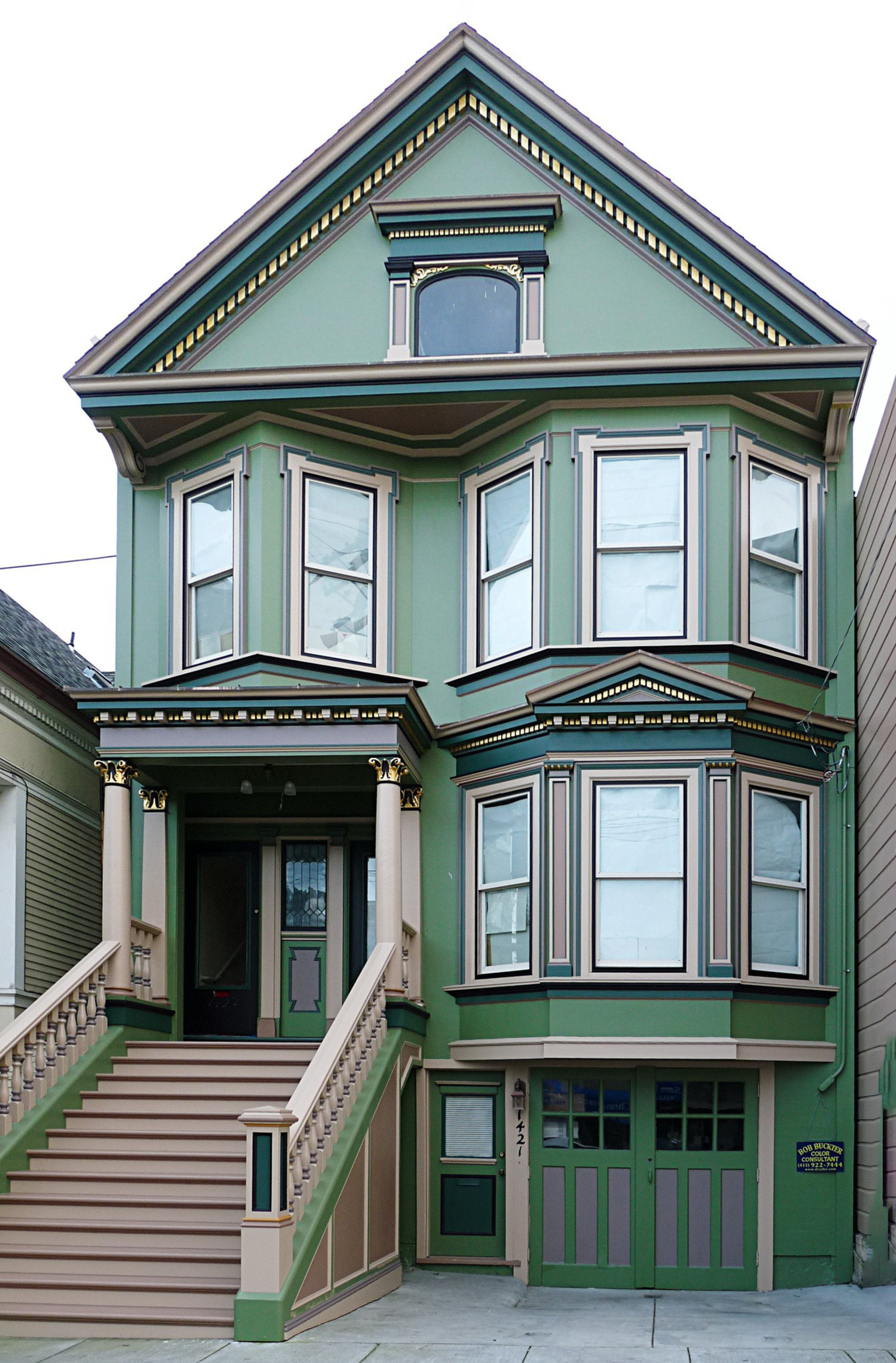A green Victorian-style house with beige trim features bay windows, a garage, and stairs leading up to a covered porch with decorative columns.