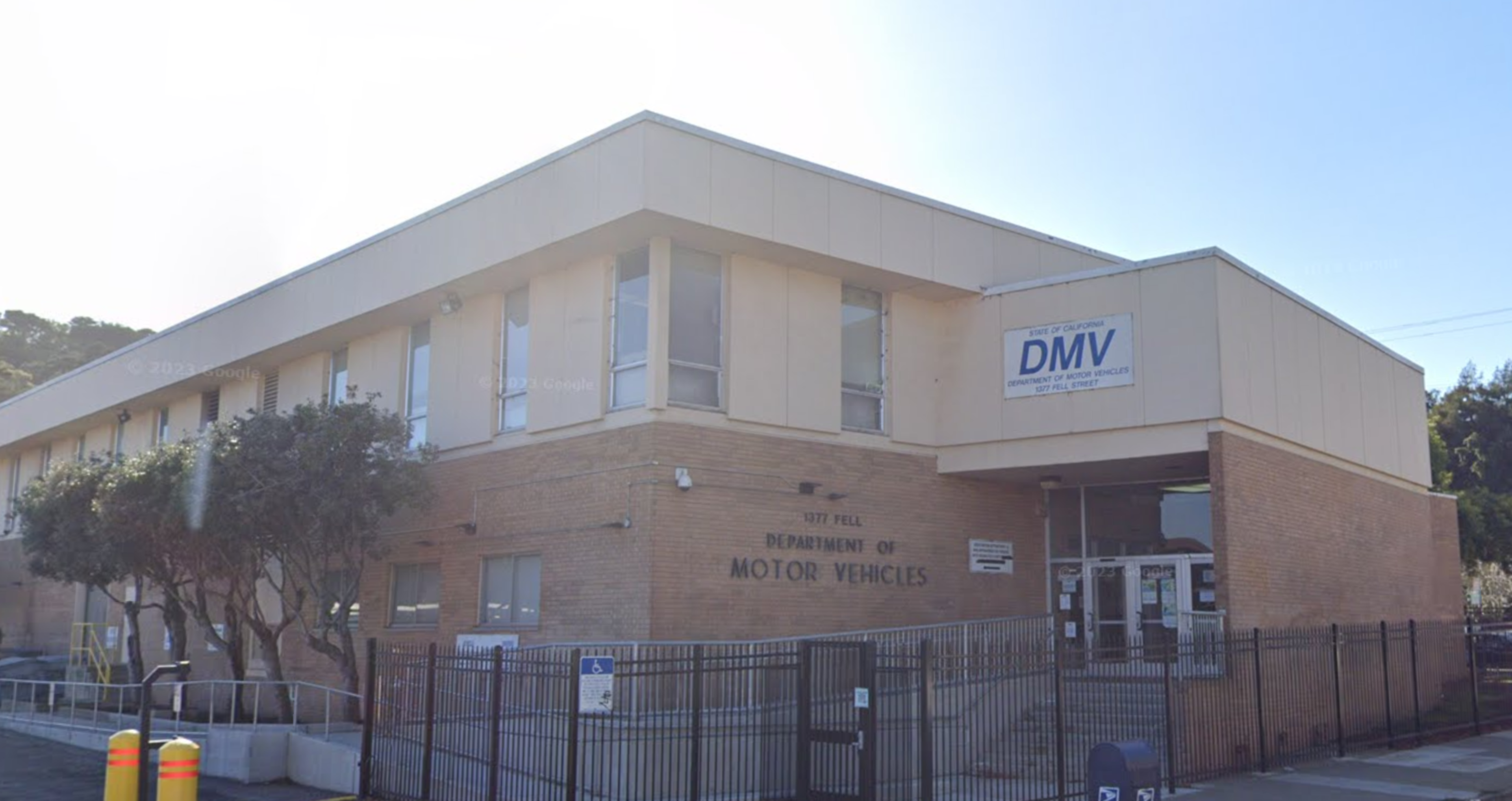 A two-story brick and beige building labeled "Department of Motor Vehicles," with a fenced entrance and a few trees along the left side.