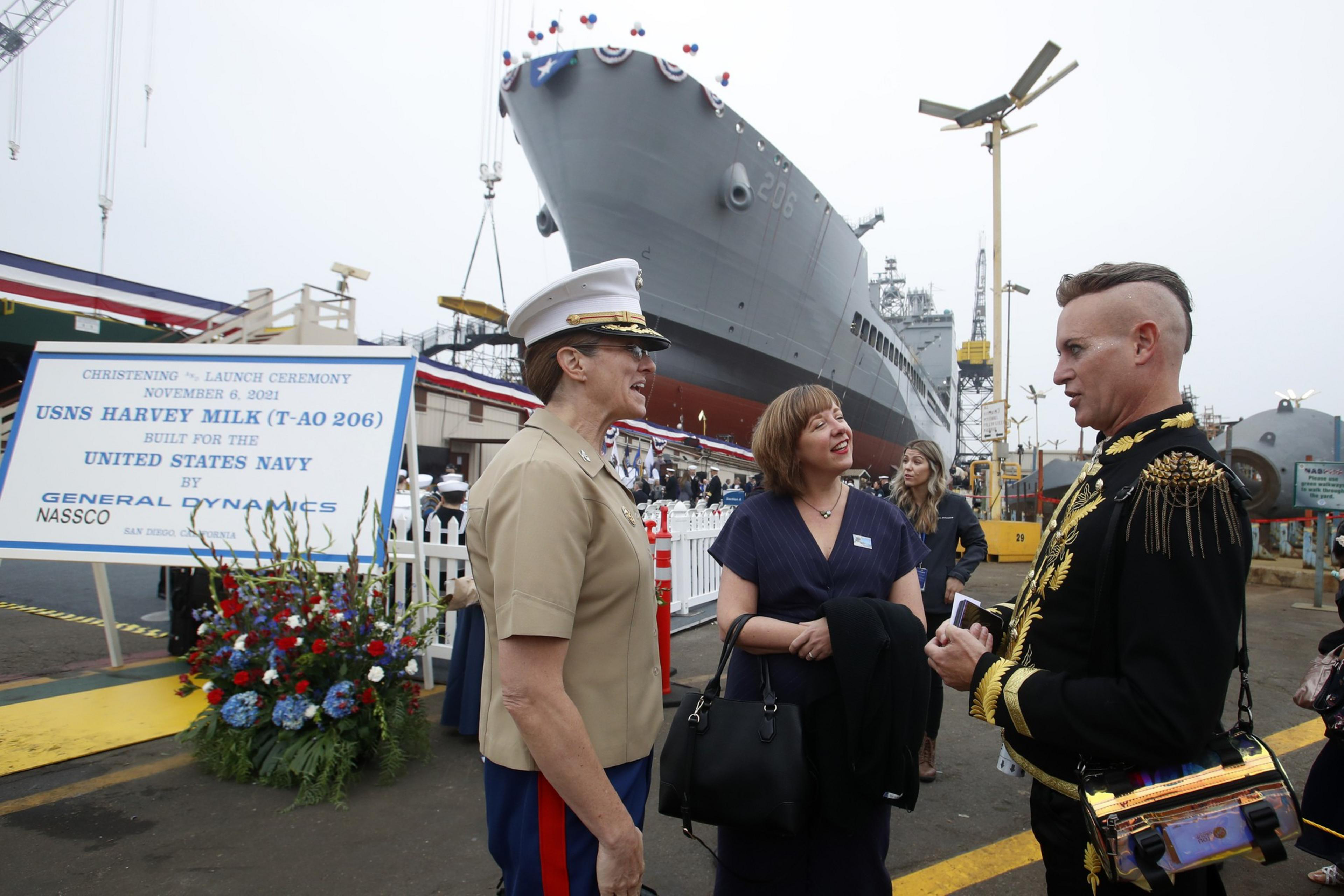 Three people converse at a ship christening event. Behind them, a large navy ship is docked. A sign indicates it's the USNS Harvey Milk launching.