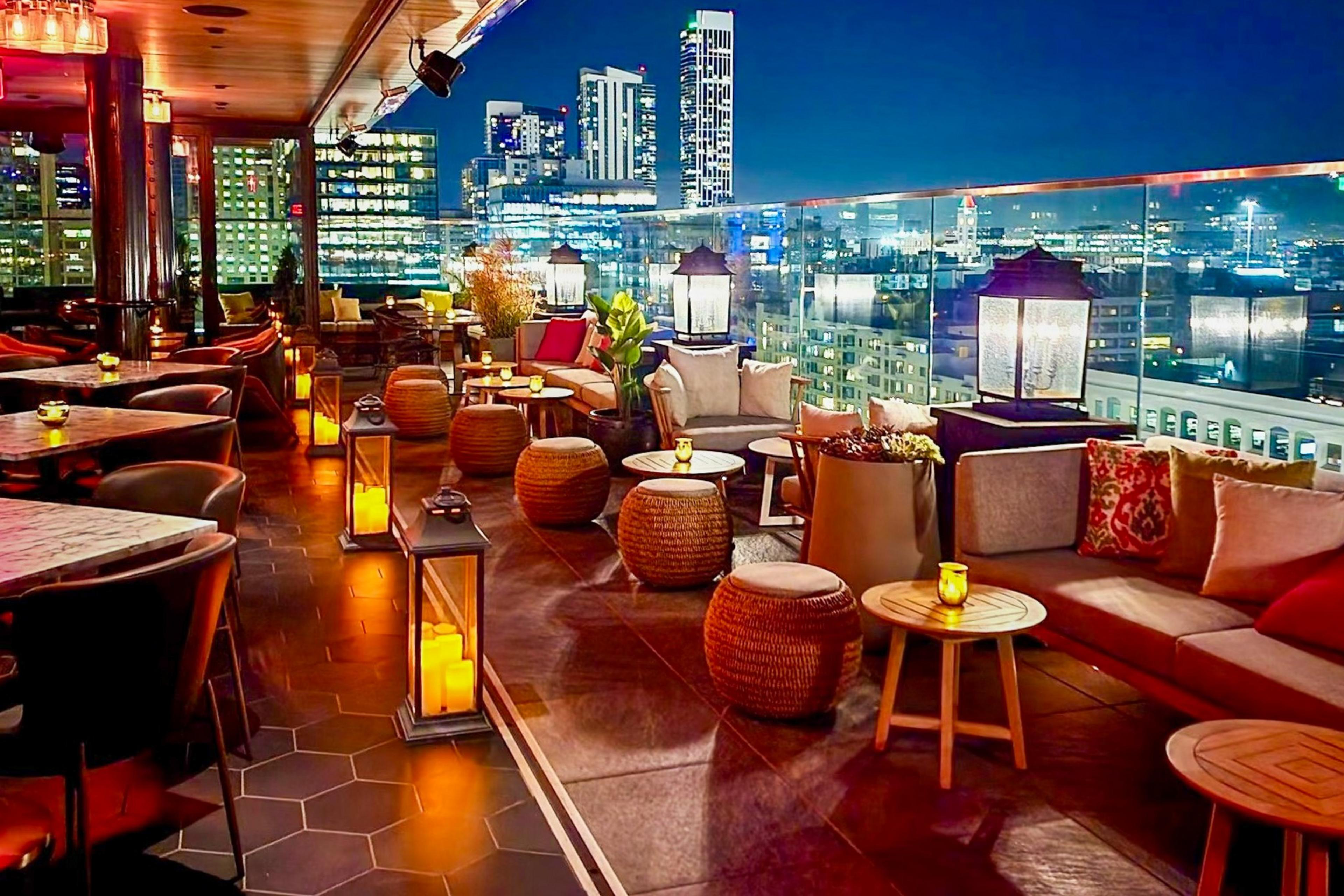A rooftop lounge at night with cushioned seating, wicker stools, lanterns with candles, small tables, plants, and city skyscrapers in the background.