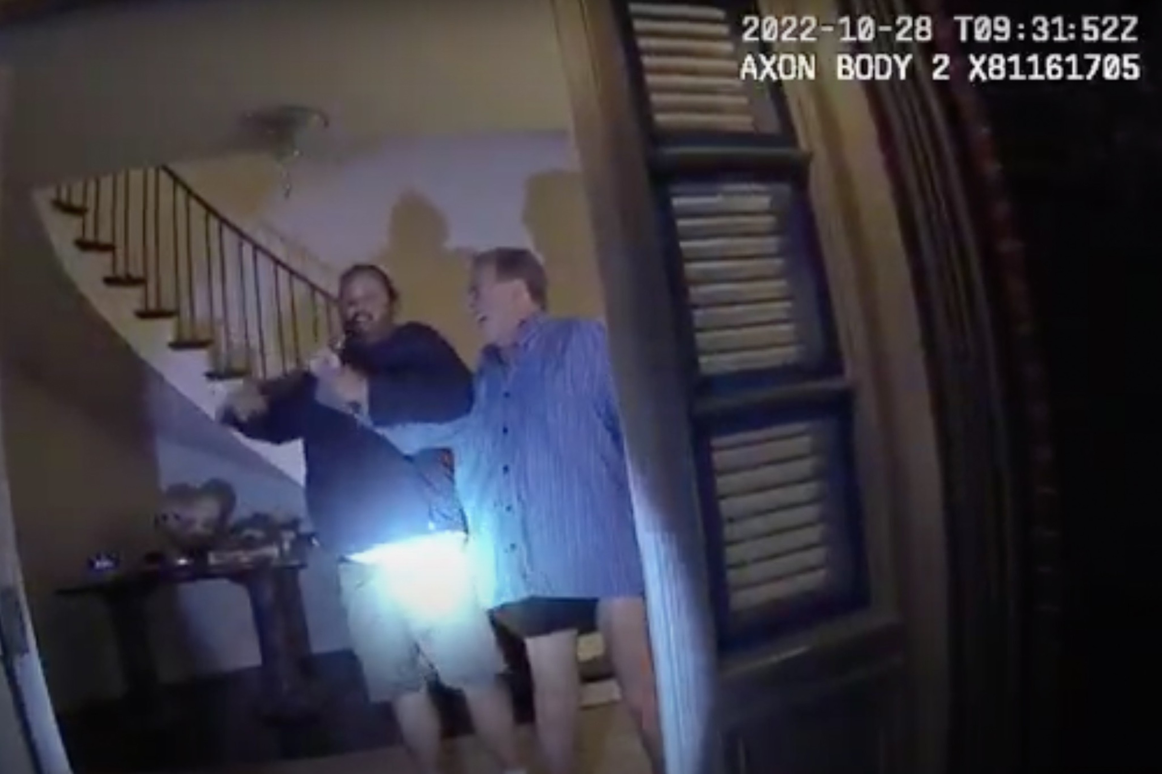 Two men stand inside an open doorway; one holds a flashlight illuminating the scene. A staircase is visible in the background.
