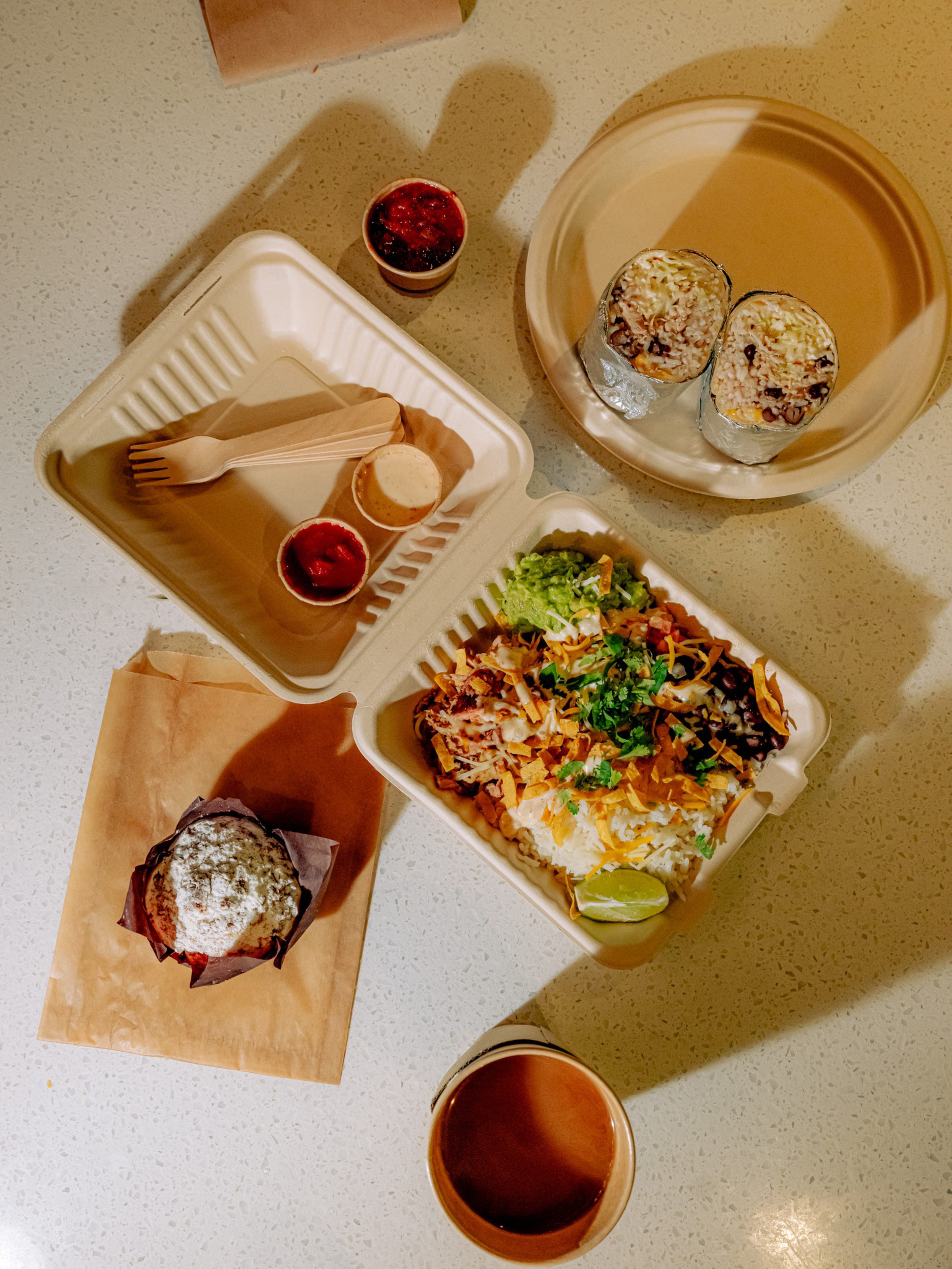 A meal spread with two burrito halves in a bowl, a container with rice, beans, lettuce, lime, sauces, a dusted muffin on paper, and a cup of coffee.
