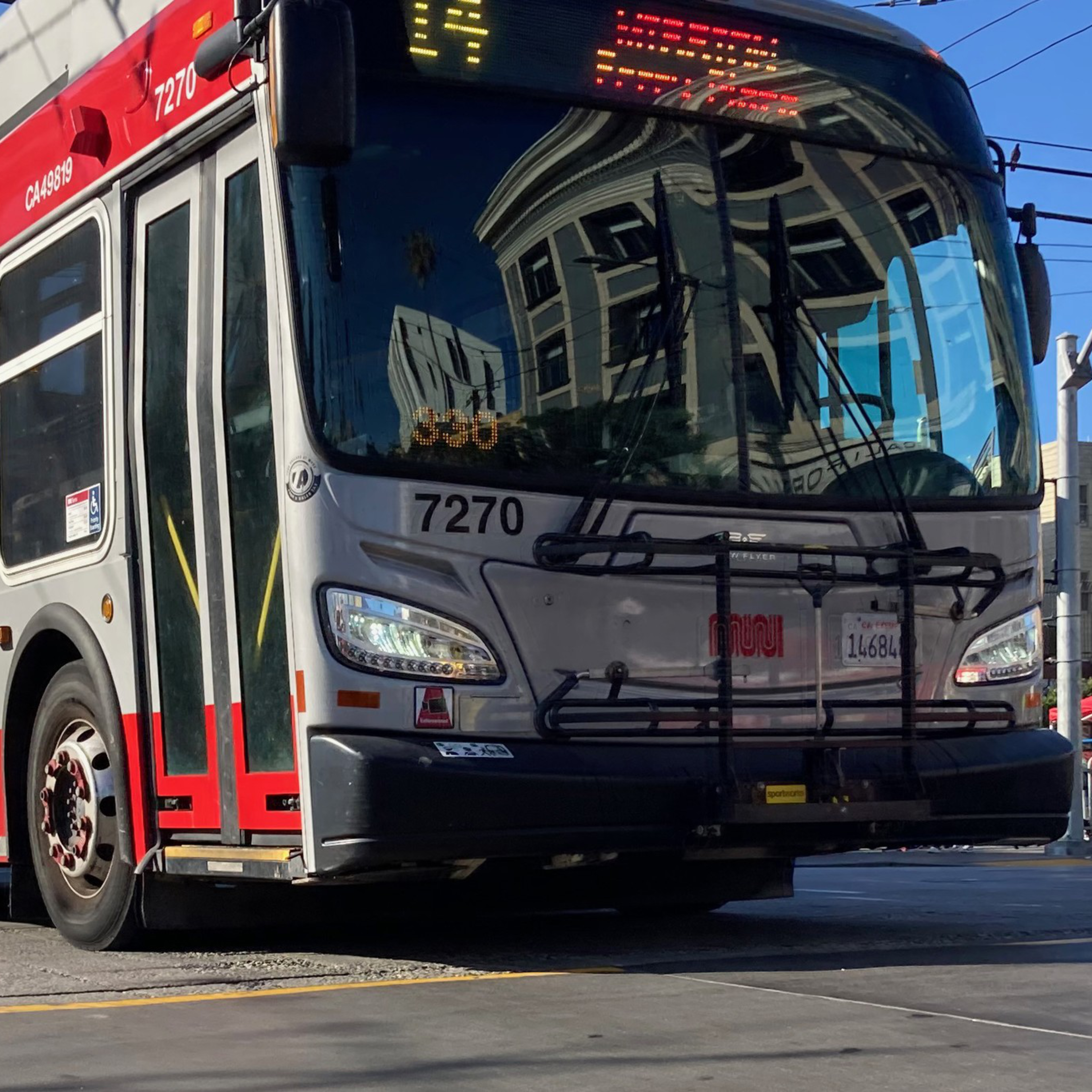 A red and gray city bus is driving on a street, with overhead wires visible above. It has the number 7270 on the front and a building reflection on its windshield.