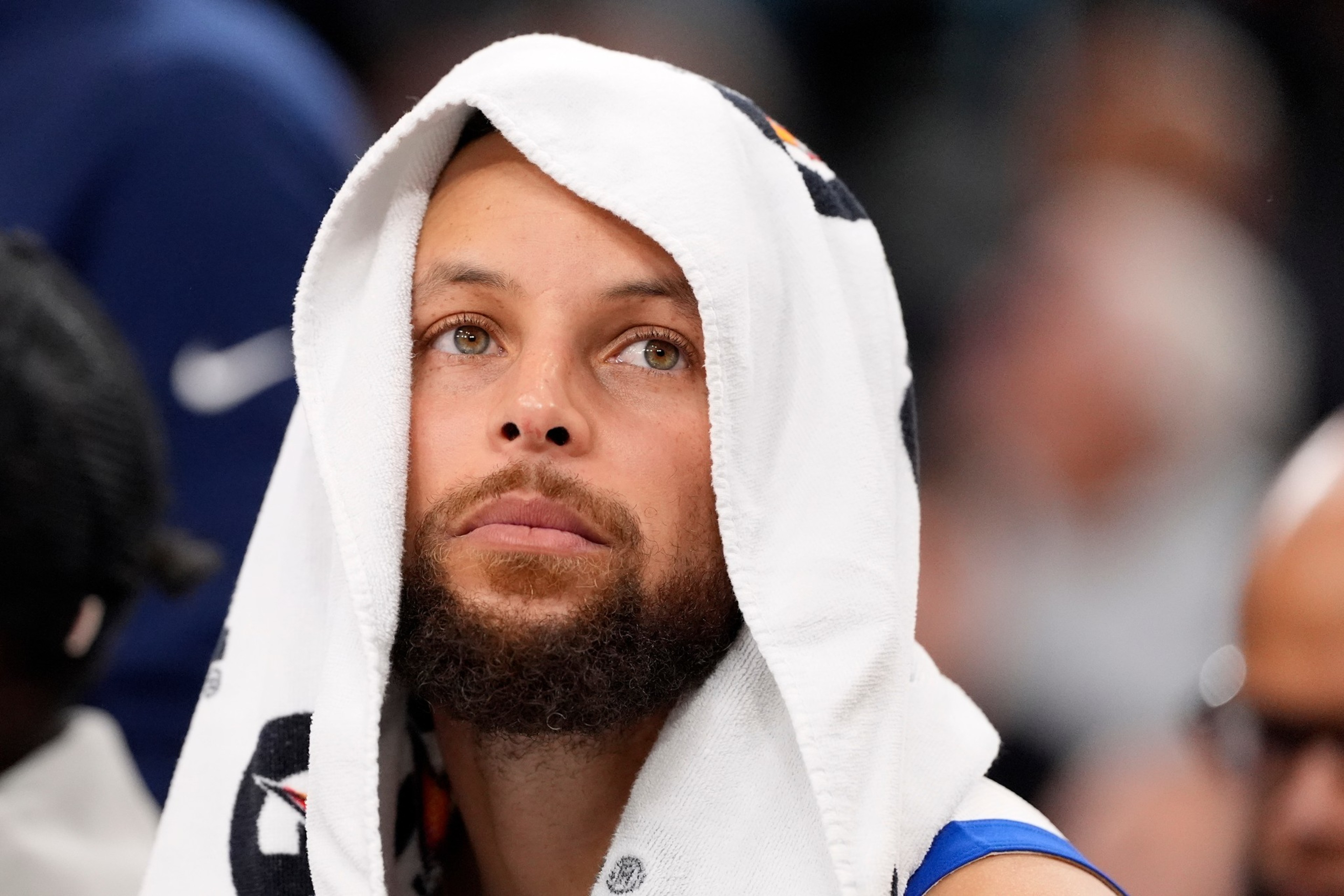A man with a beard and light brown eyes has a white towel draped over his head, looking thoughtful and slightly upward.