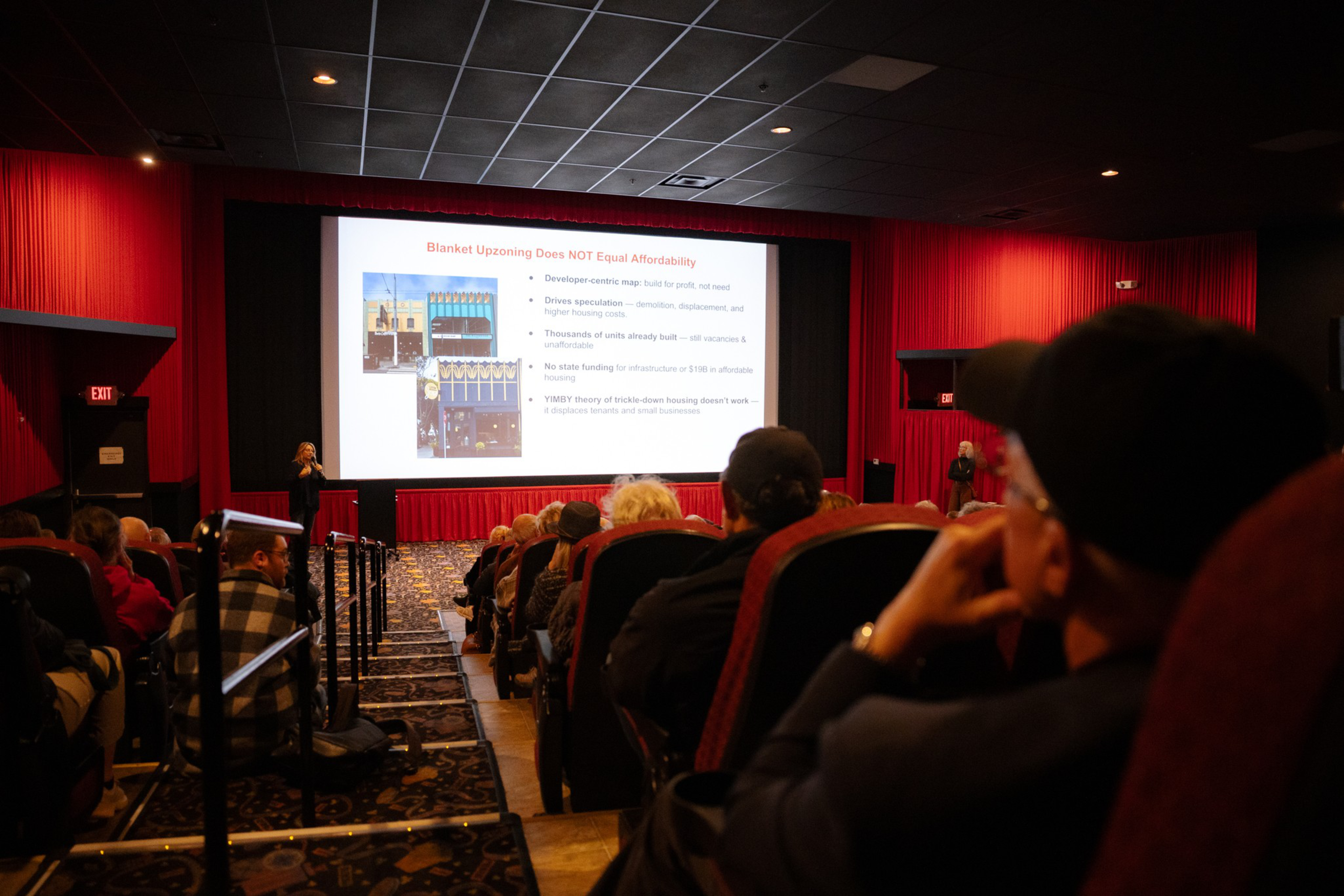 A theater room with red curtains, showing a presentation titled &quot;Blanket Upzoning Does NOT Equal Affordability.&quot; People are seated, listening attentively.