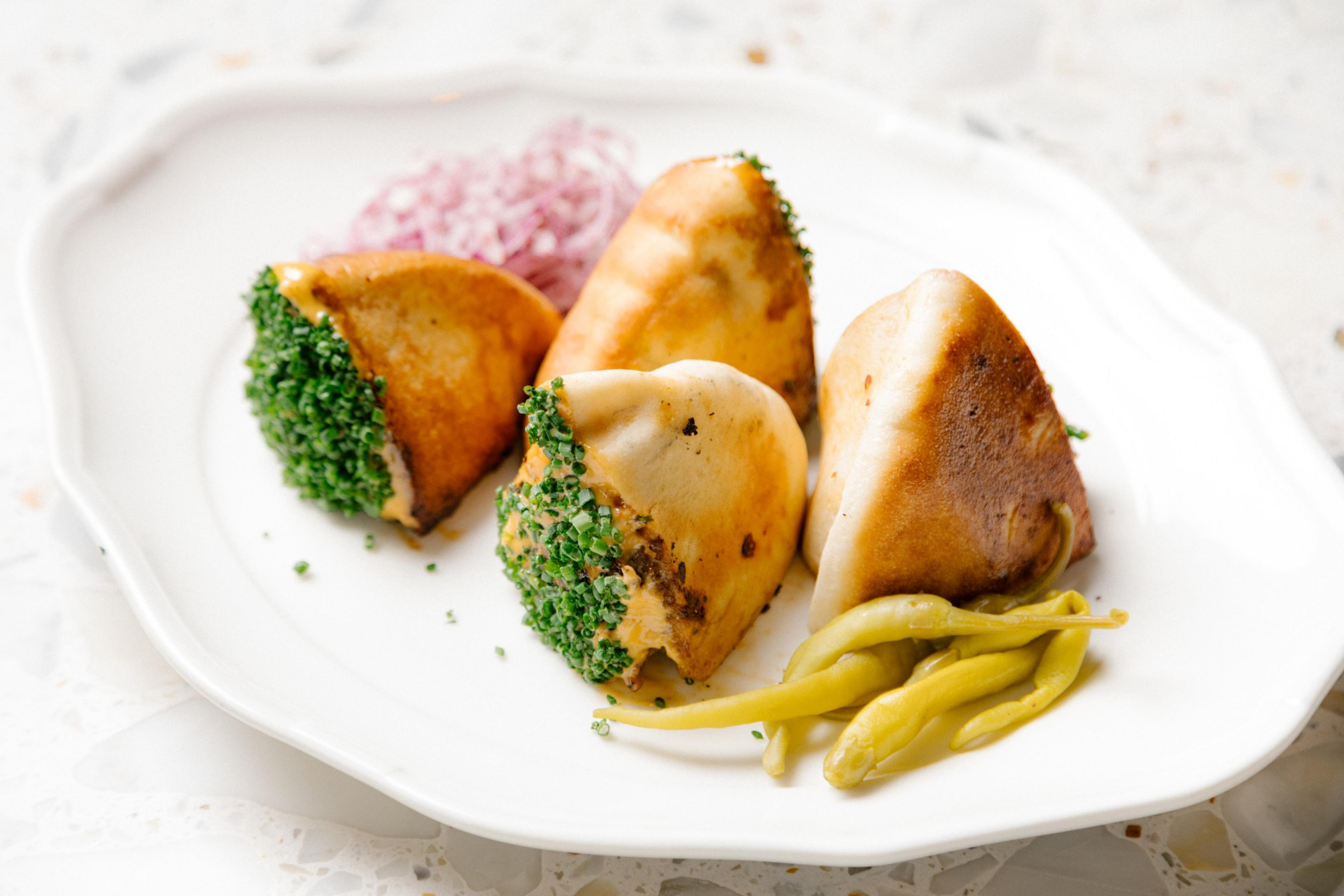 A close up 4 pastry like looking item on a white ceramic plate that are the &quot;Arayes Burger&quot; dish and include the ingredients meat kebab cooked in a pita, shatta emulsion, chives, guindilla.