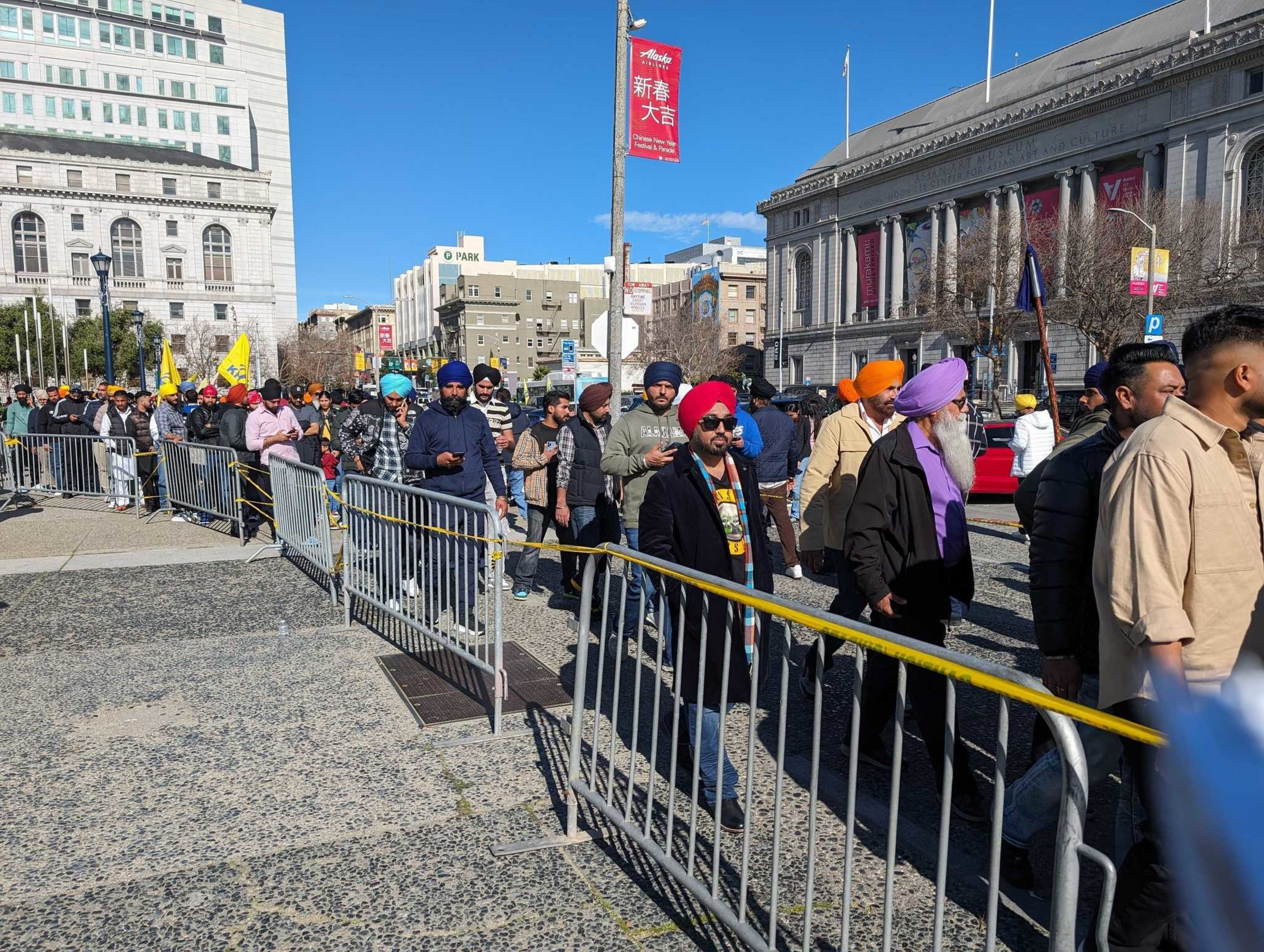 A line of people in casual clothing standing beside security fencing in a public plaza