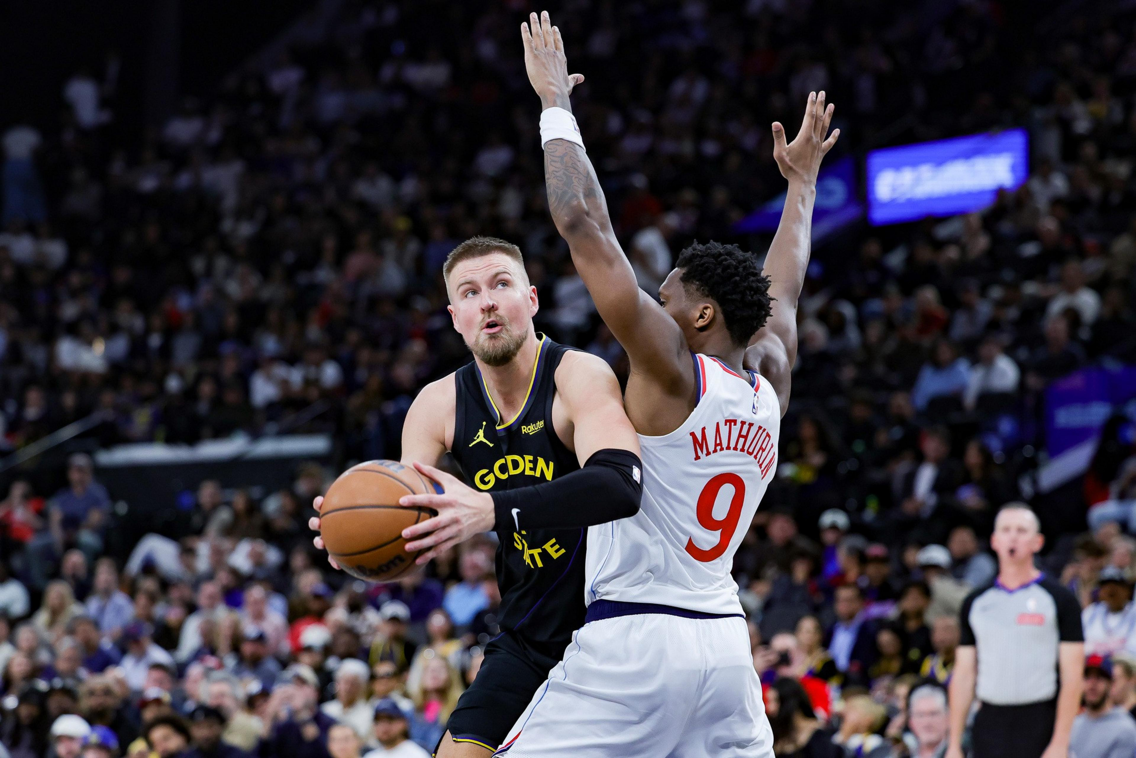 A Golden State Warriors player holds the basketball, preparing to shoot while a Philadelphia 76ers player with arms raised defends him.