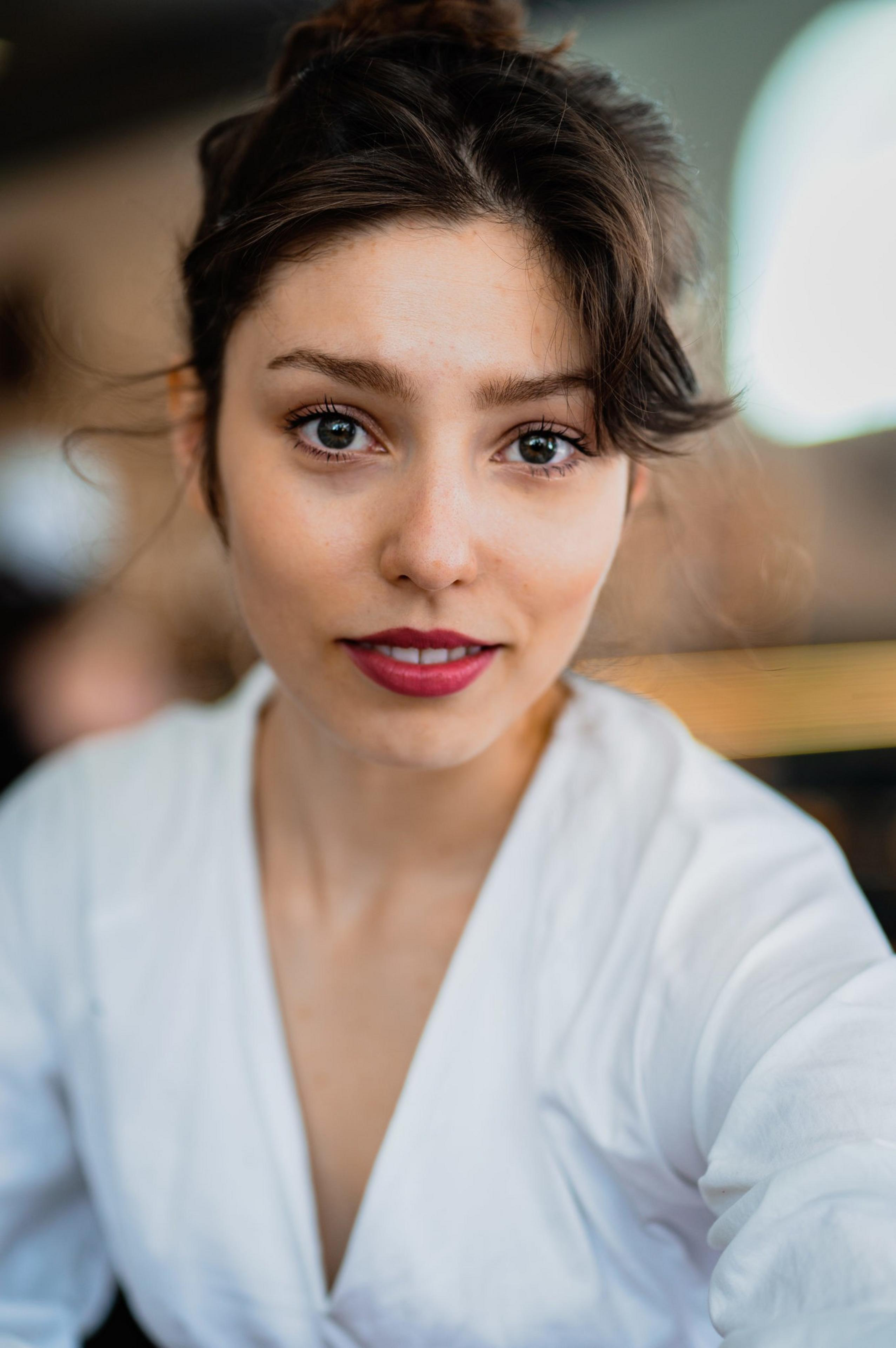 A person with brown hair and red lipstick is smiling softly, wearing a white shirt, with a blurred background giving a cozy and warm feel.