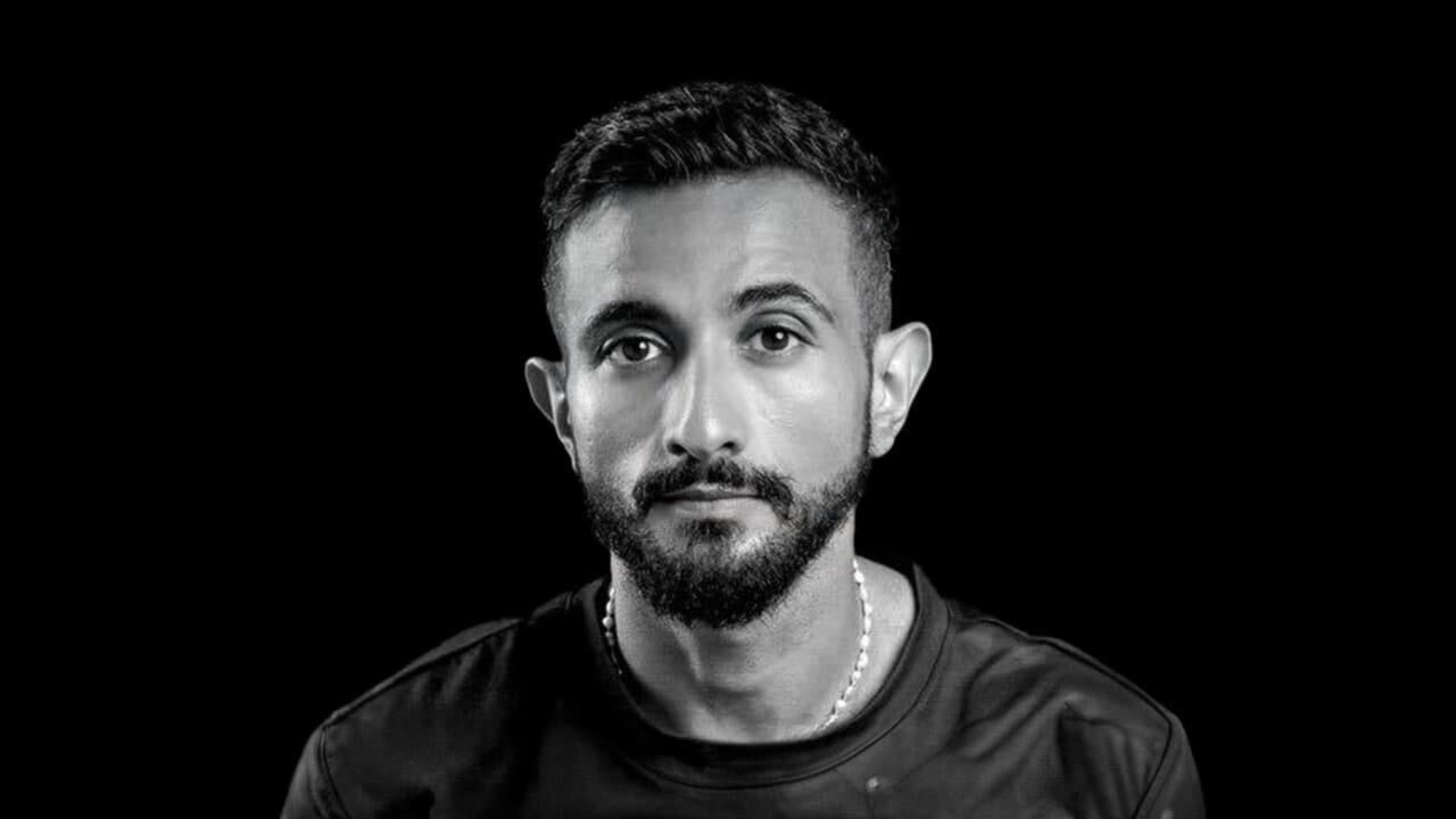 A man with short, dark hair and a trimmed beard looks directly ahead, wearing a dark shirt and a thin beaded necklace, against a black background.