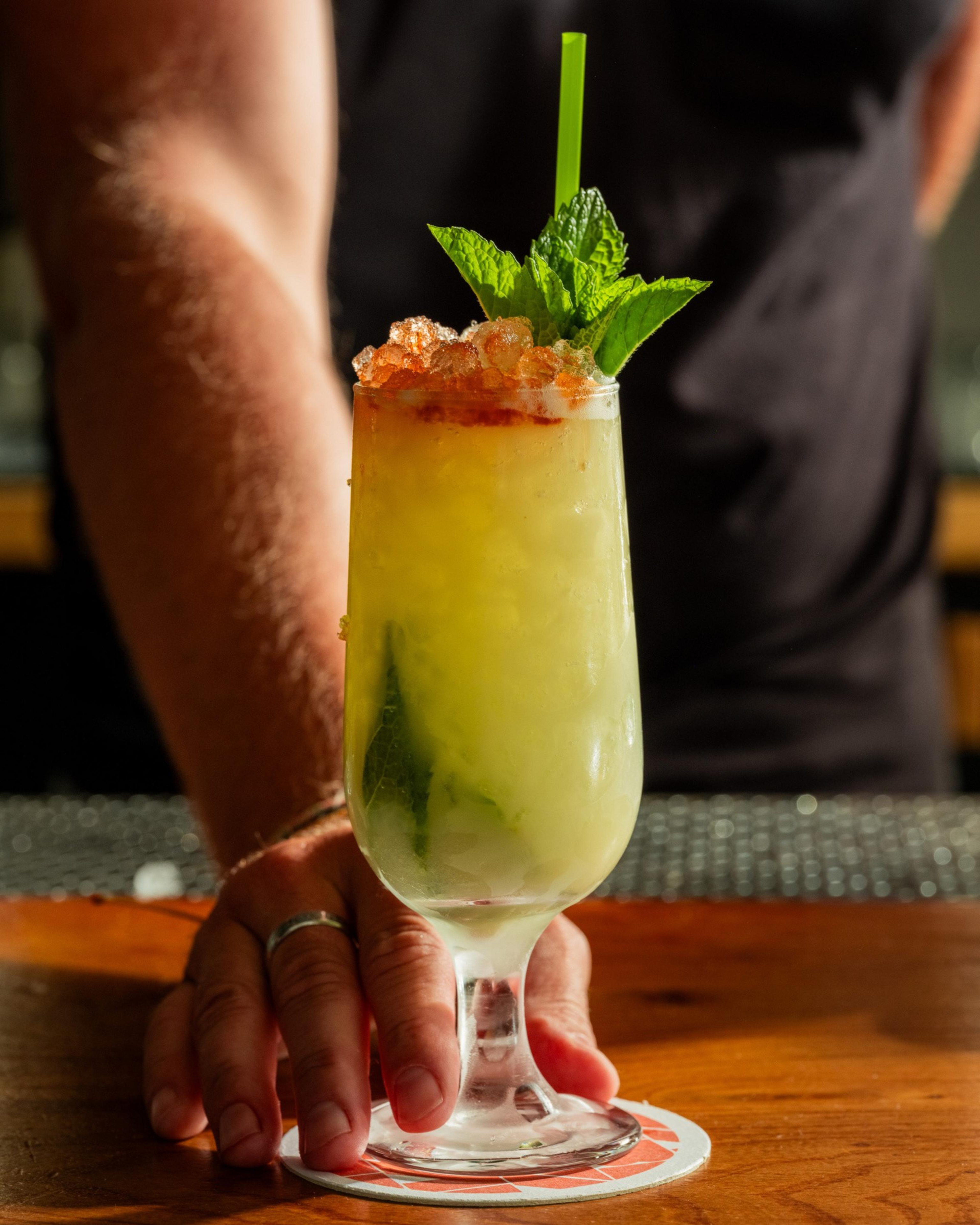 A frosty cocktail in a glass adorned with crushed ice, mint leaves, and a green straw sits on a coaster, held by a hand.