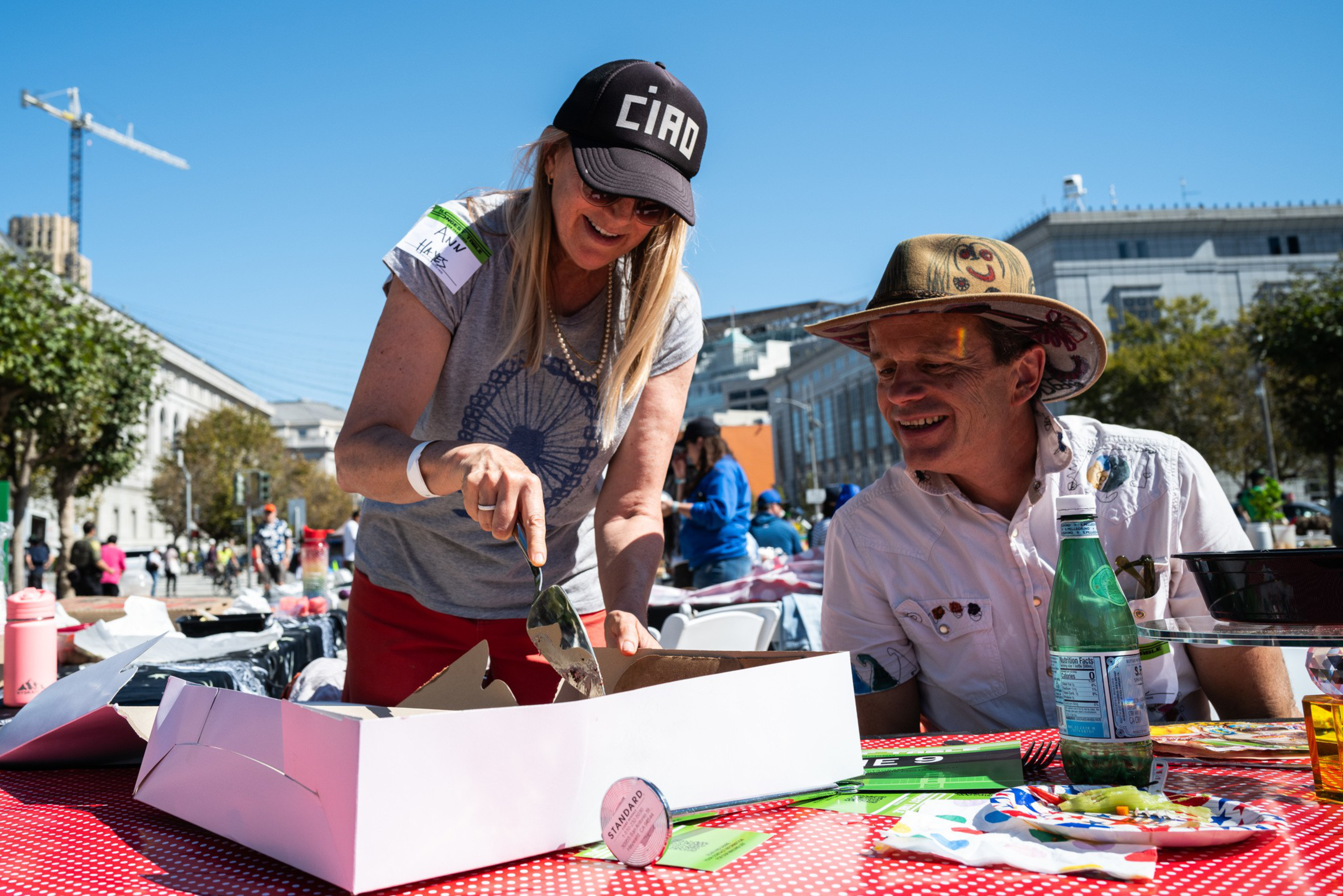 A woman wearing a “CIAO” cap and sunglasses cuts pizza while a smiling man in a hat watches at a sunny outdoor event with tables and people in the background.