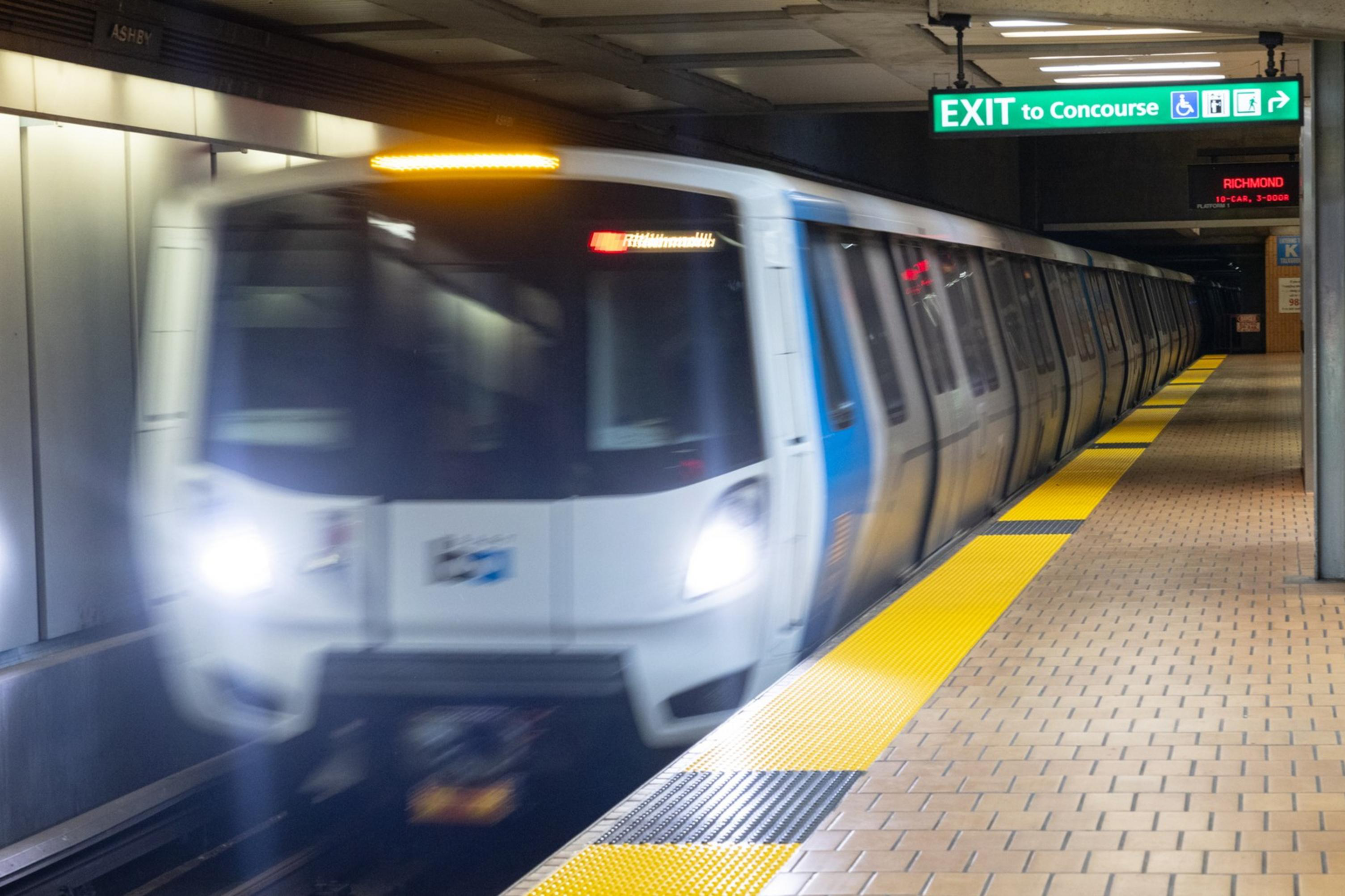 A subway train approaches the Ashby station as an elderly woman, wearing sunglasses and a green jacket, checks her phone beside a black suitcase near an "Exit to Concourse" sign.