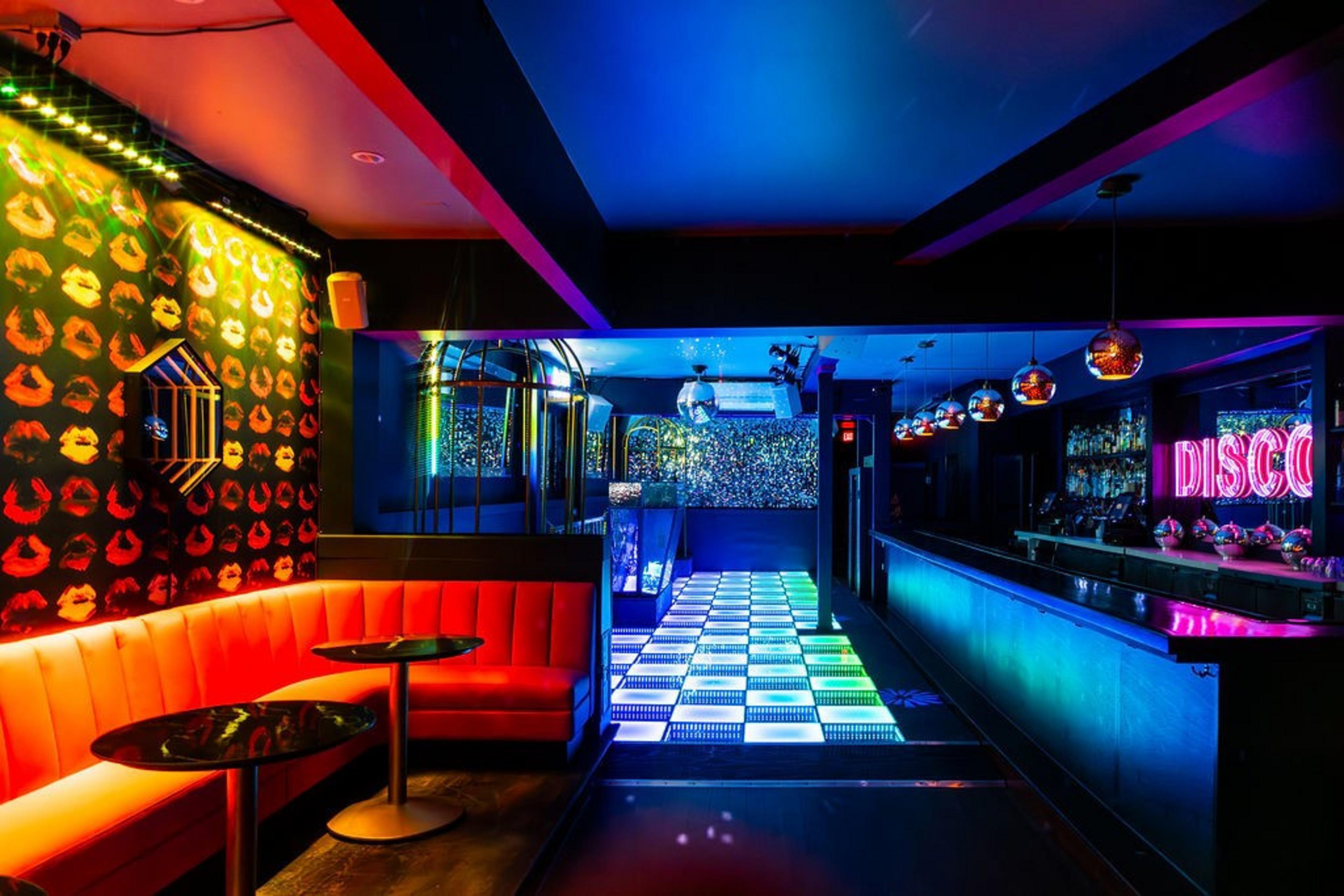 A vibrant, colorful nightclub with a light-up dance floor, neon &quot;DISCO&quot; sign, red seating, walls adorned with lip prints, and a long, illuminated bar.