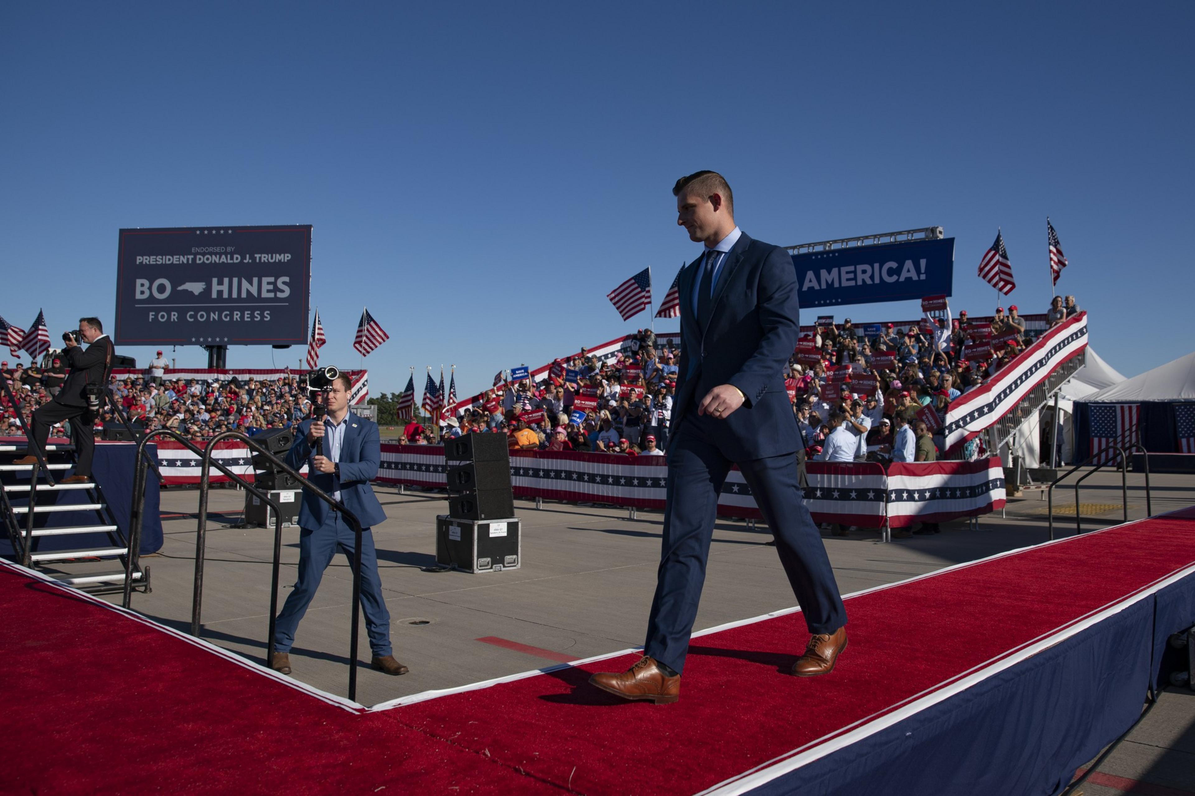 A man in a blue suit walks on a red carpeted stage at a rally. The crowd, holding American flags, is seated in bleachers under a clear blue sky.