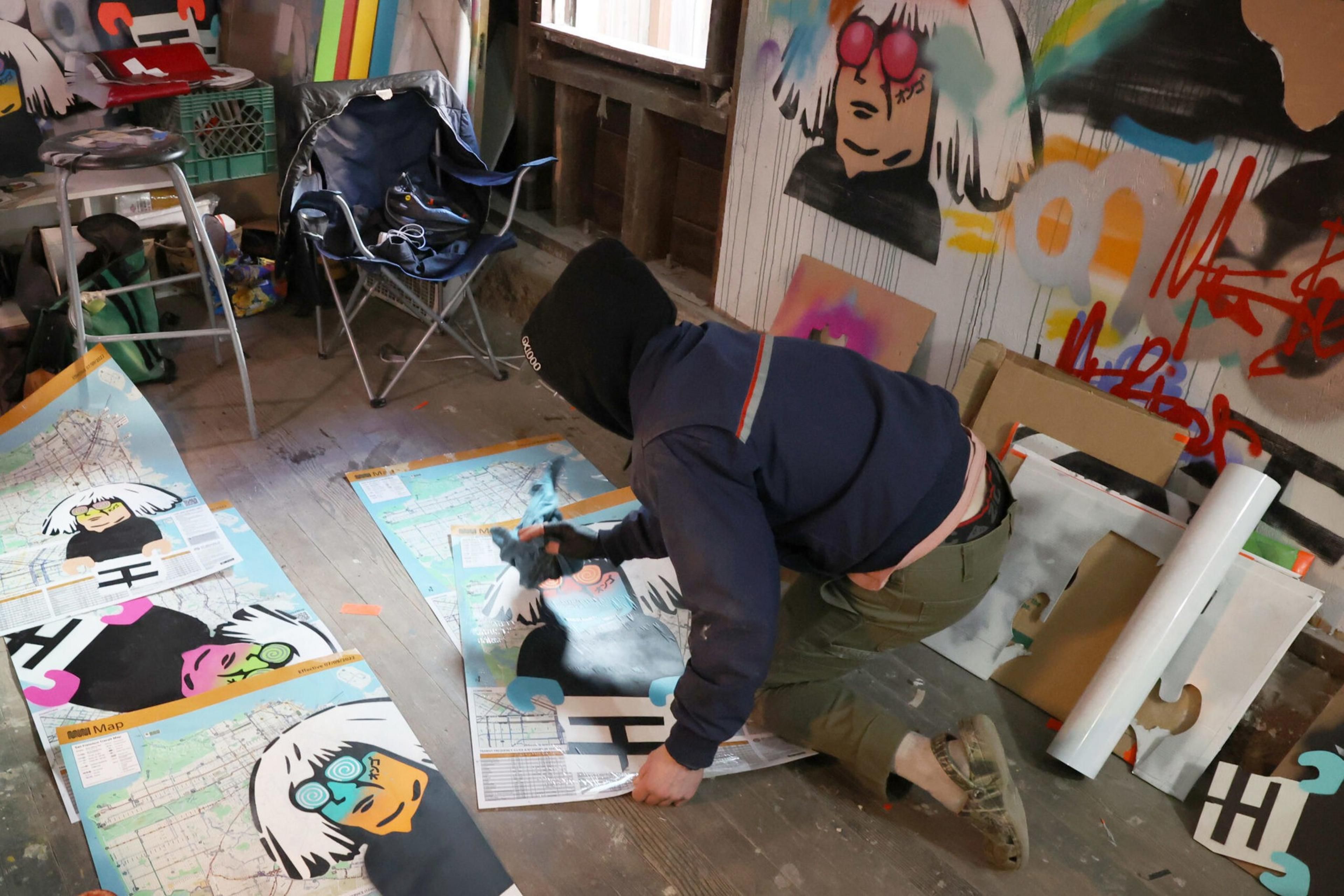 A person wearing a hoodie and gloves sprays paint over large street maps with cartoon faces while kneeling on a wooden floor in an art-filled room.