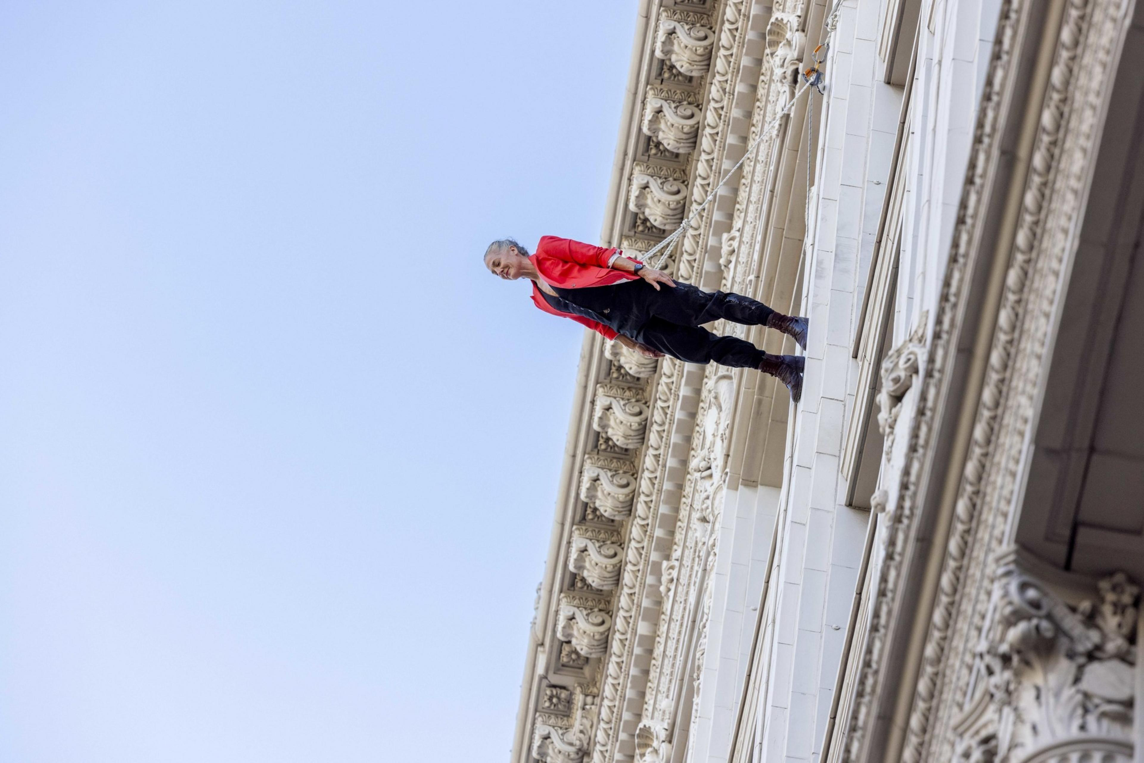 A person in a red jacket and black pants is suspended horizontally on the side of an ornate building, seemingly defying gravity against a clear blue sky.
