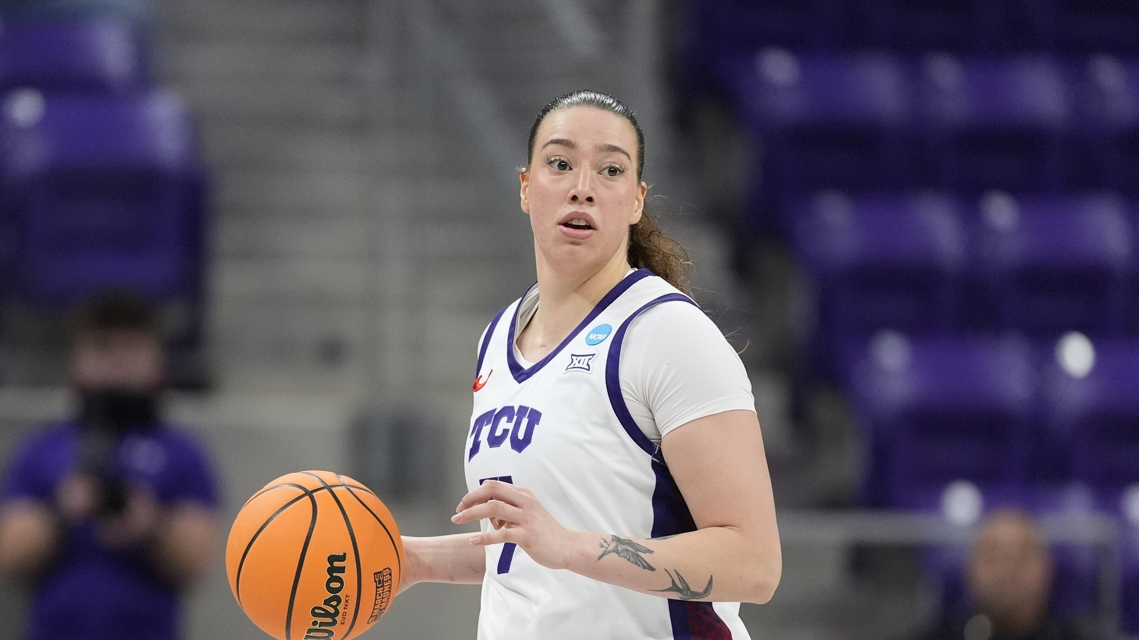 Una jugadora de baloncesto con una camiseta blanca de TCU está driblando una pelota de baloncesto Wilson naranja en una cancha con espectadores borrosos en el fondo.