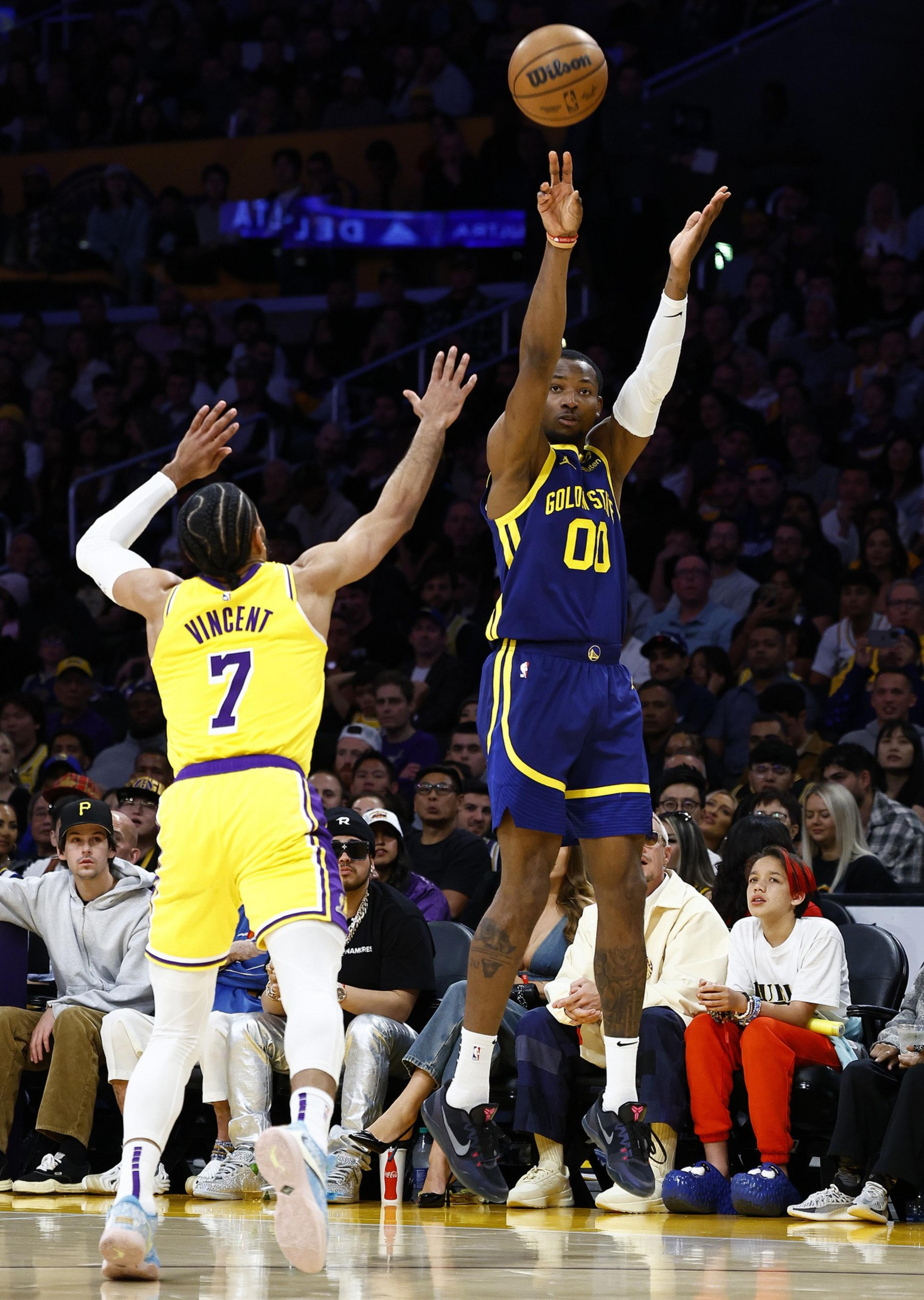 A basketball player in a blue &quot;Golden State&quot; jersey is shooting a jump shot, while a player in a yellow &quot;7&quot; jersey tries to block. The crowd watches intently.