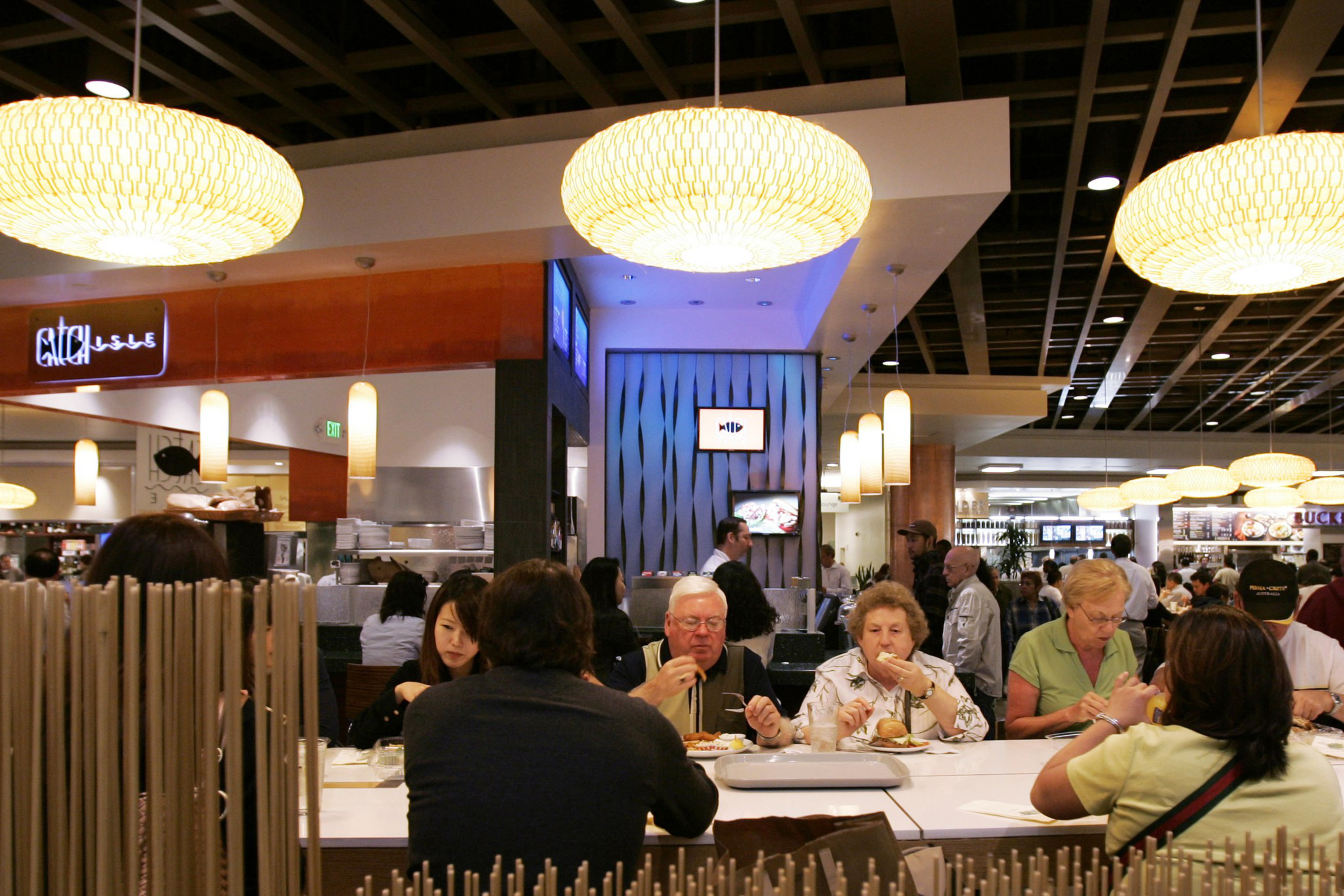 A busy food court with diners at tables, large hanging lamps, and a variety of food stalls in the background.