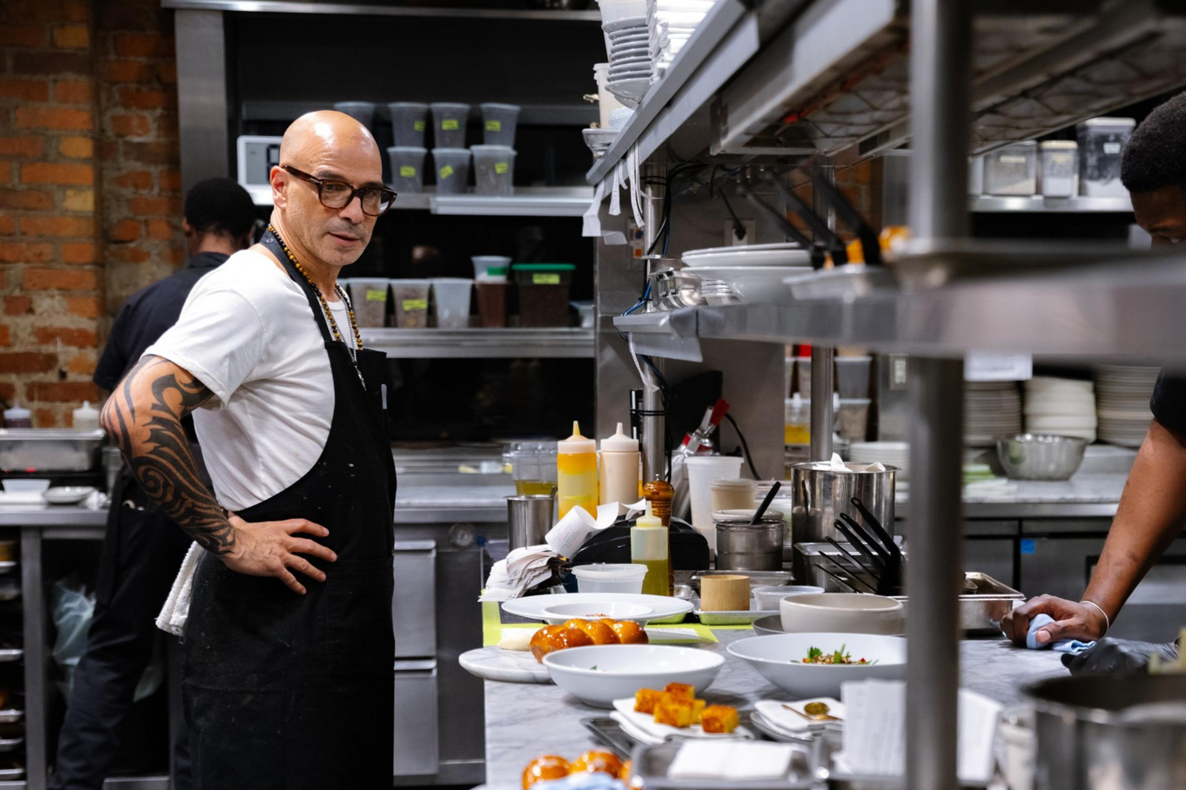 Two chefs work in a bustling kitchen; one stands with hands on hips, the other prepares food at a counter. The space is filled with utensils and ingredients.