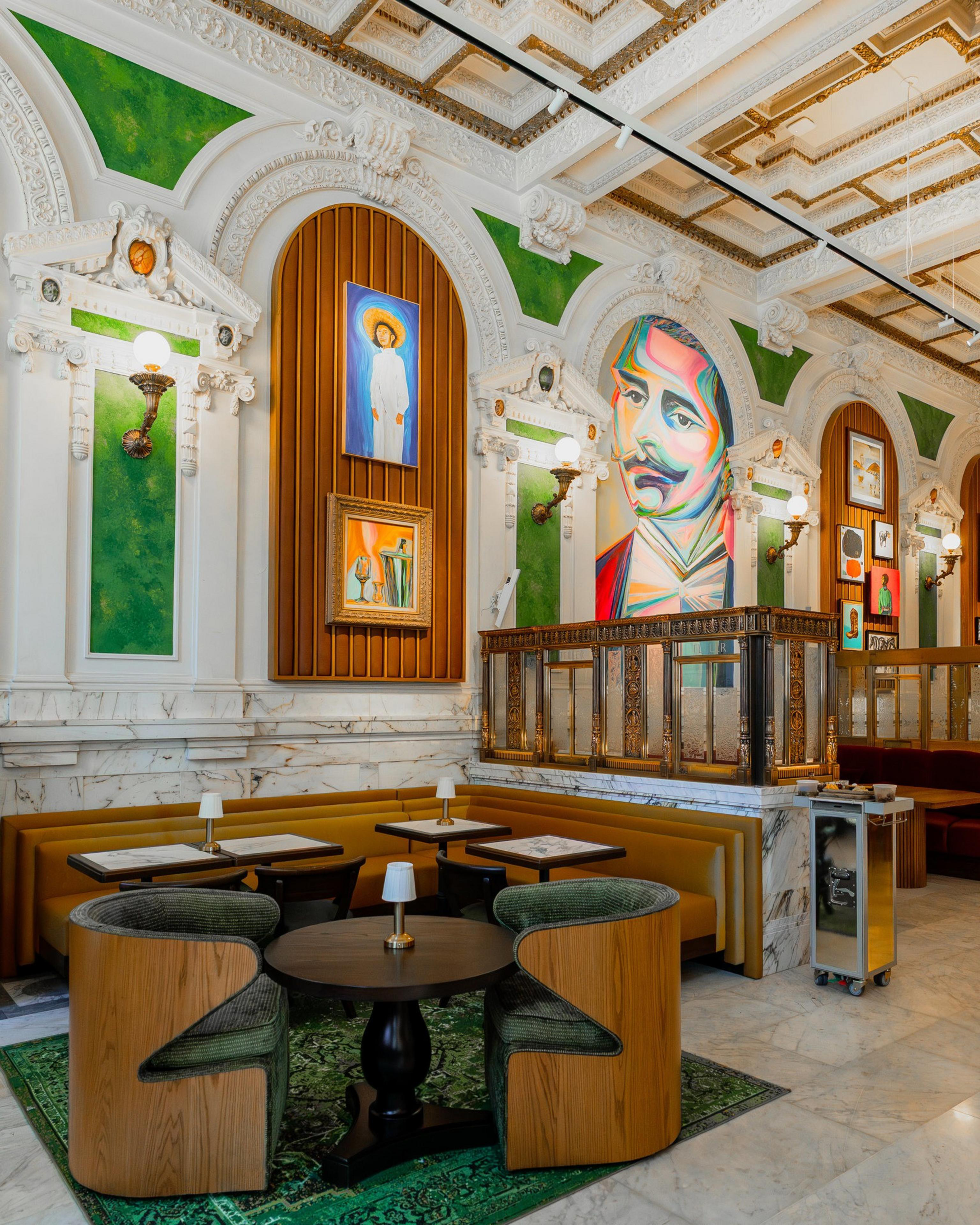 A stylish cafe with green velvet chairs, wooden tables, ornate white walls with green accents, and colorful modern portraits hangs above a marble half-wall.