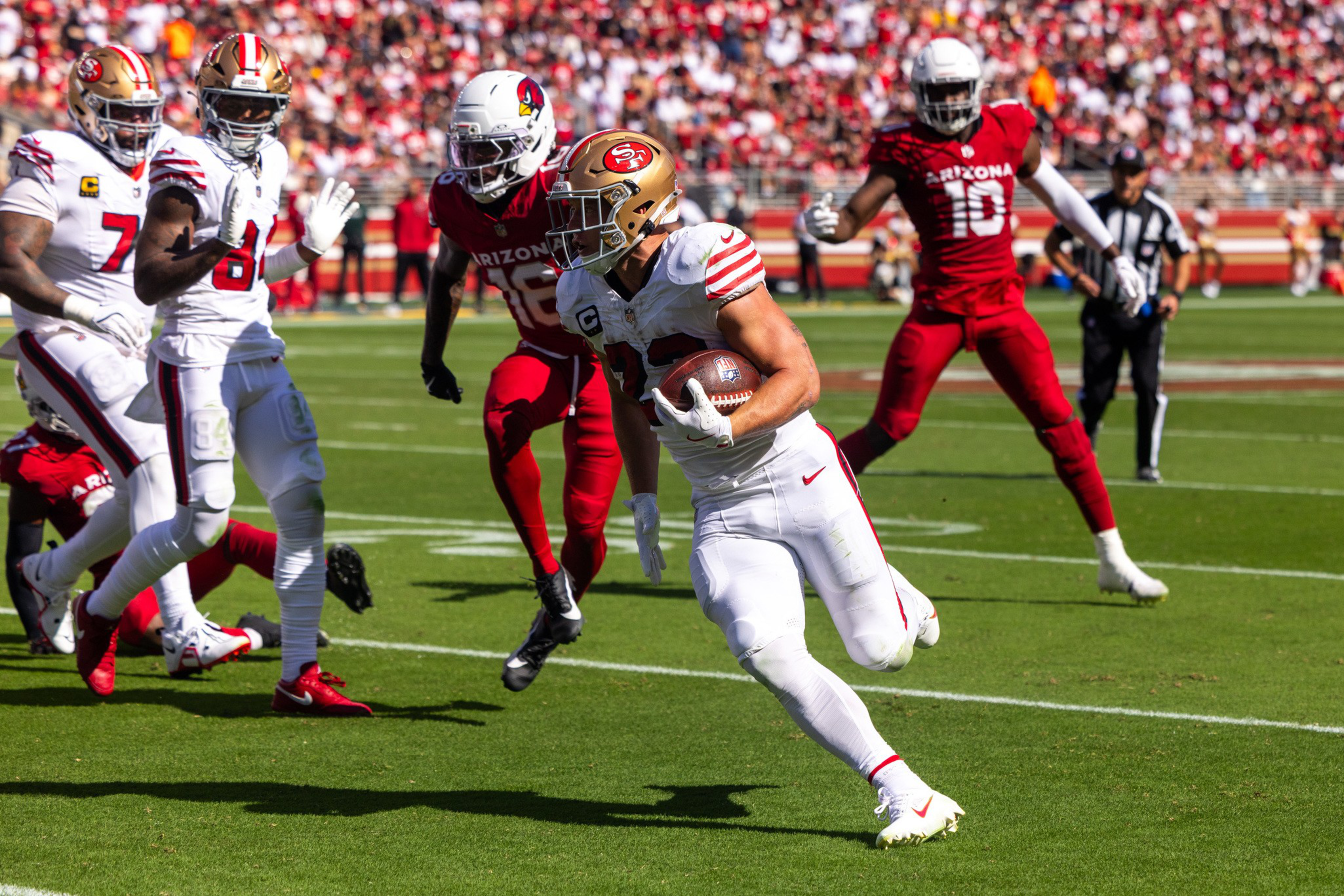 A football player in a white uniform runs with the ball while players in red uniforms chase him on a sunny field.