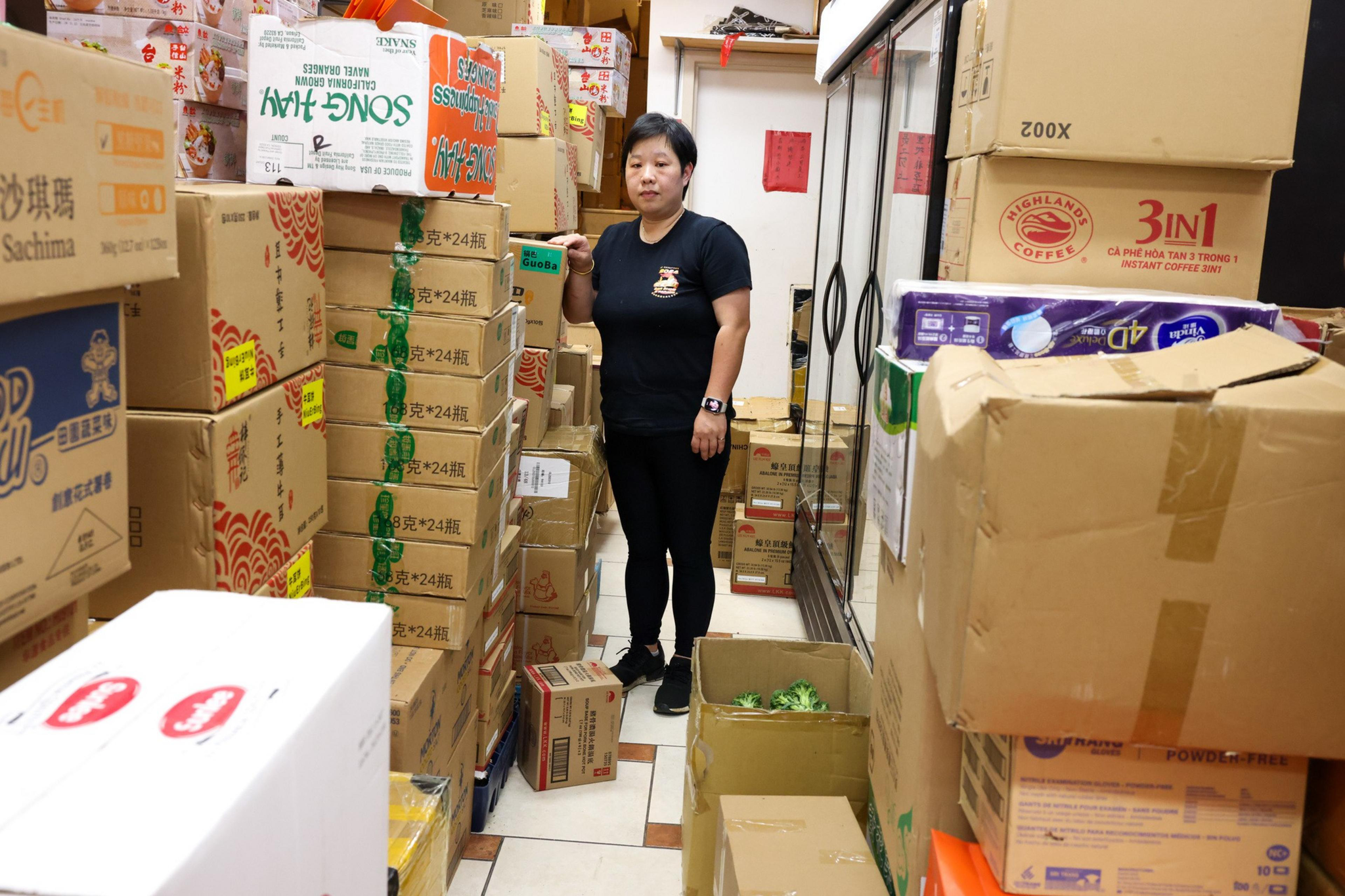 A woman stands in a narrow storage area filled with stacked cardboard boxes, many labeled with Asian brands. She wears black attire and a smartwatch.