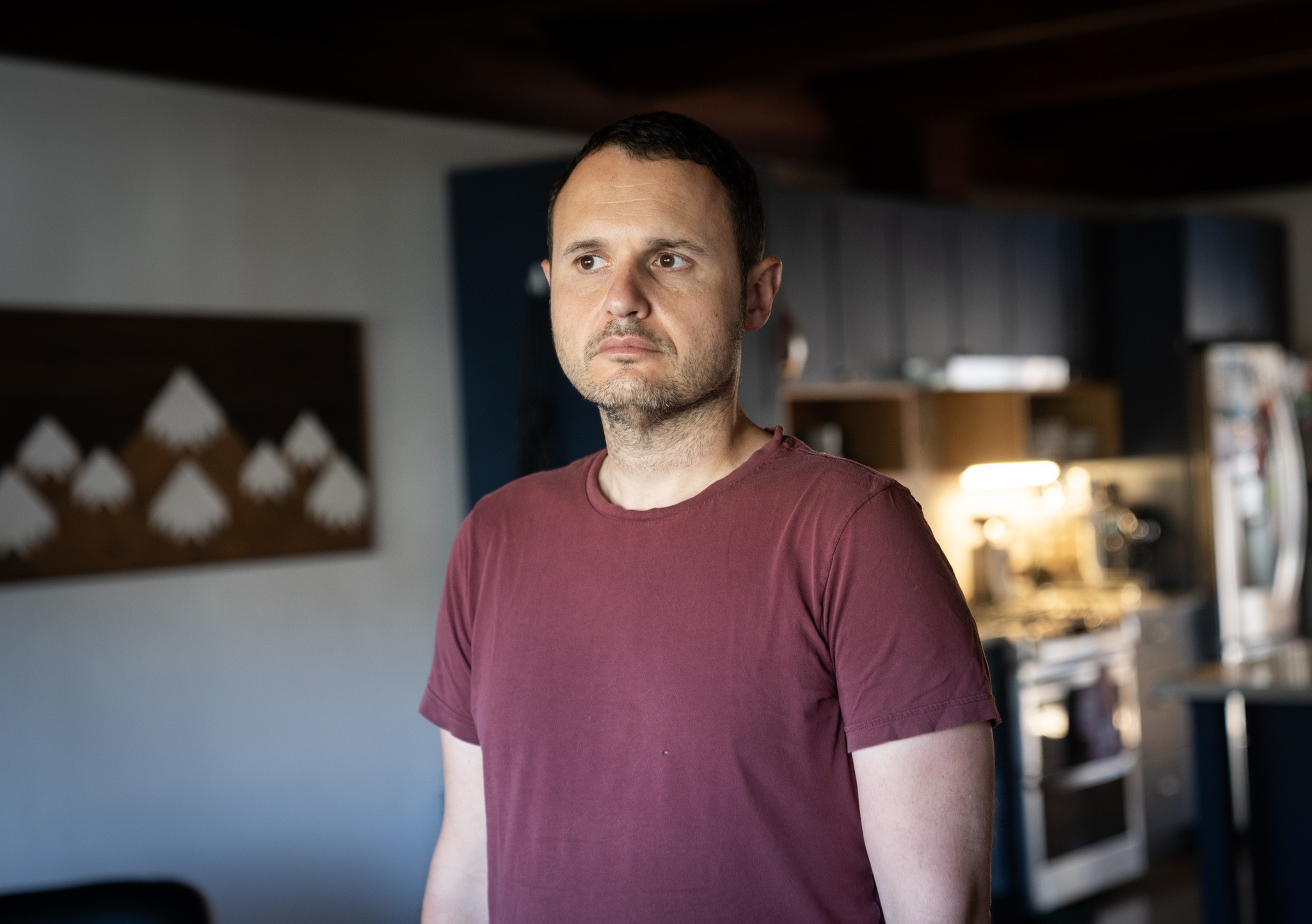 A man in a maroon t-shirt stands indoors, looking slightly to the side with a neutral expression, against a background of a kitchen and wall art.
